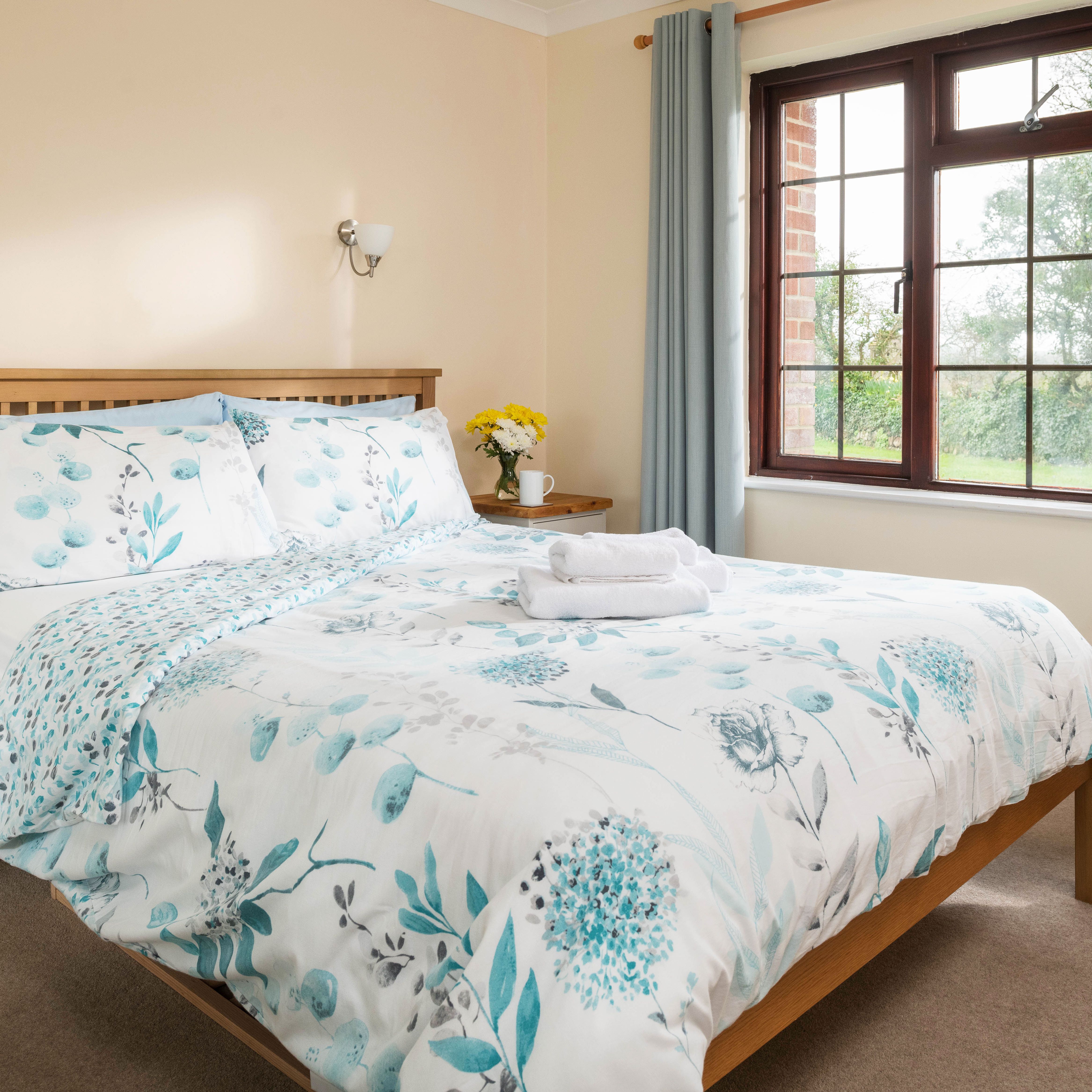 Bright and cozy bedroom with a wooden bed, floral bedding, bedside tables, a large window with blue curtains, and a chair with clothes draped over it.