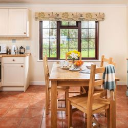 Bright, cozy kitchen with cream cabinets, wooden countertops, a wooden dining table set for breakfast, large window with garden view, tiled floor, and a sideboard with fresh flowers.