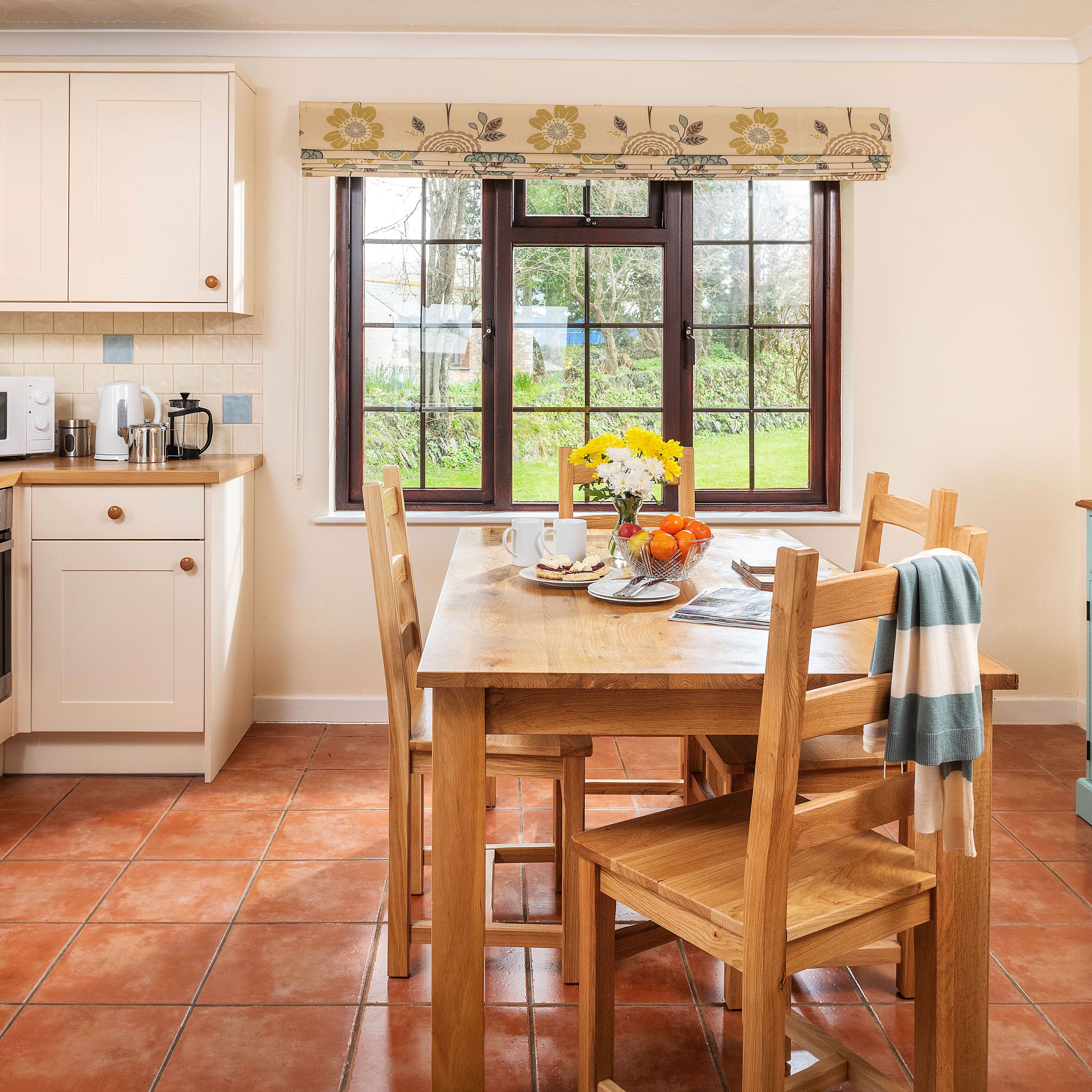 Bright, cozy kitchen with cream cabinets, wooden countertops, a wooden dining table set for breakfast, large window with garden view, tiled floor, and a sideboard with fresh flowers.