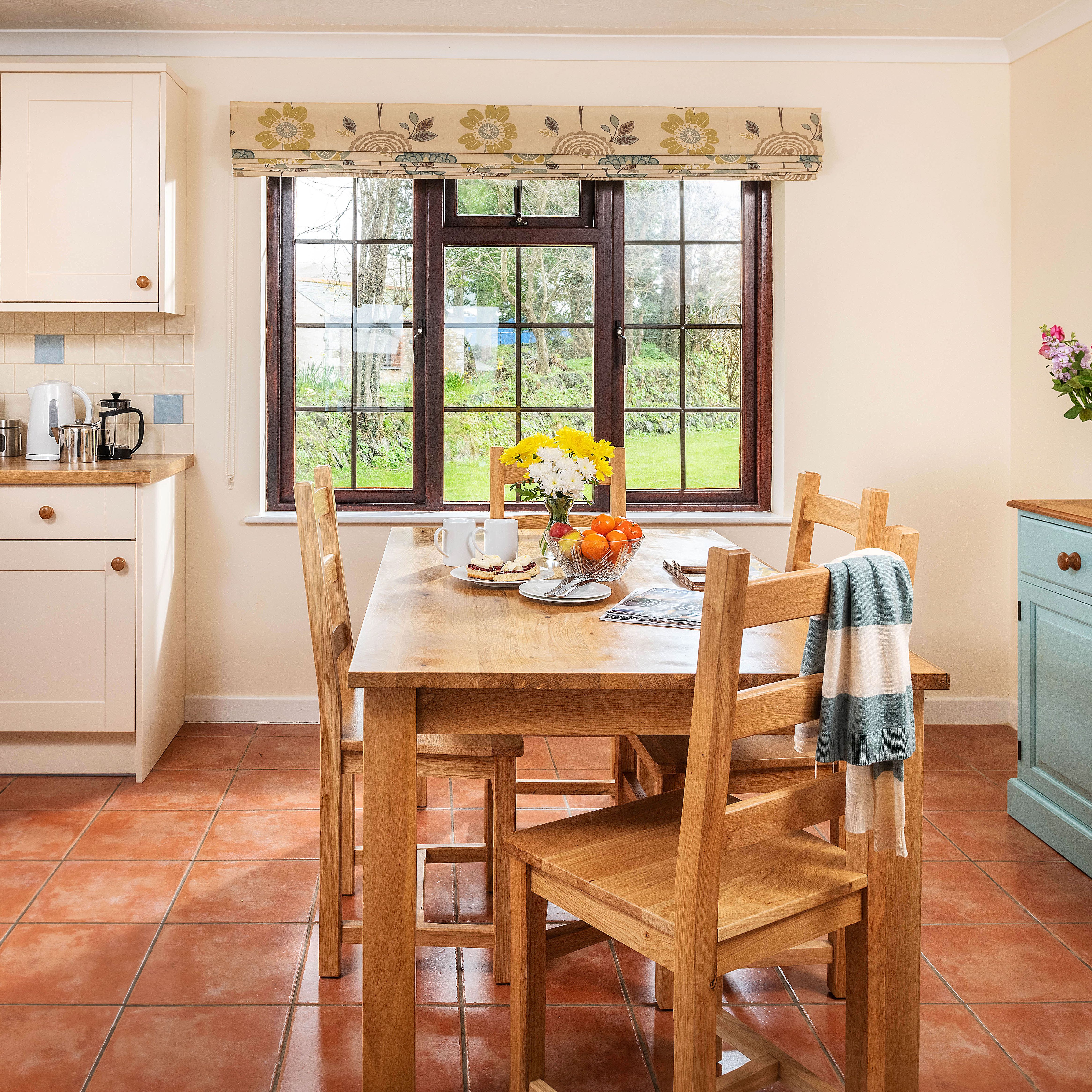 Bright, cozy kitchen with cream cabinets, wooden countertops, a wooden dining table set for breakfast, large window with garden view, tiled floor, and a sideboard with fresh flowers.