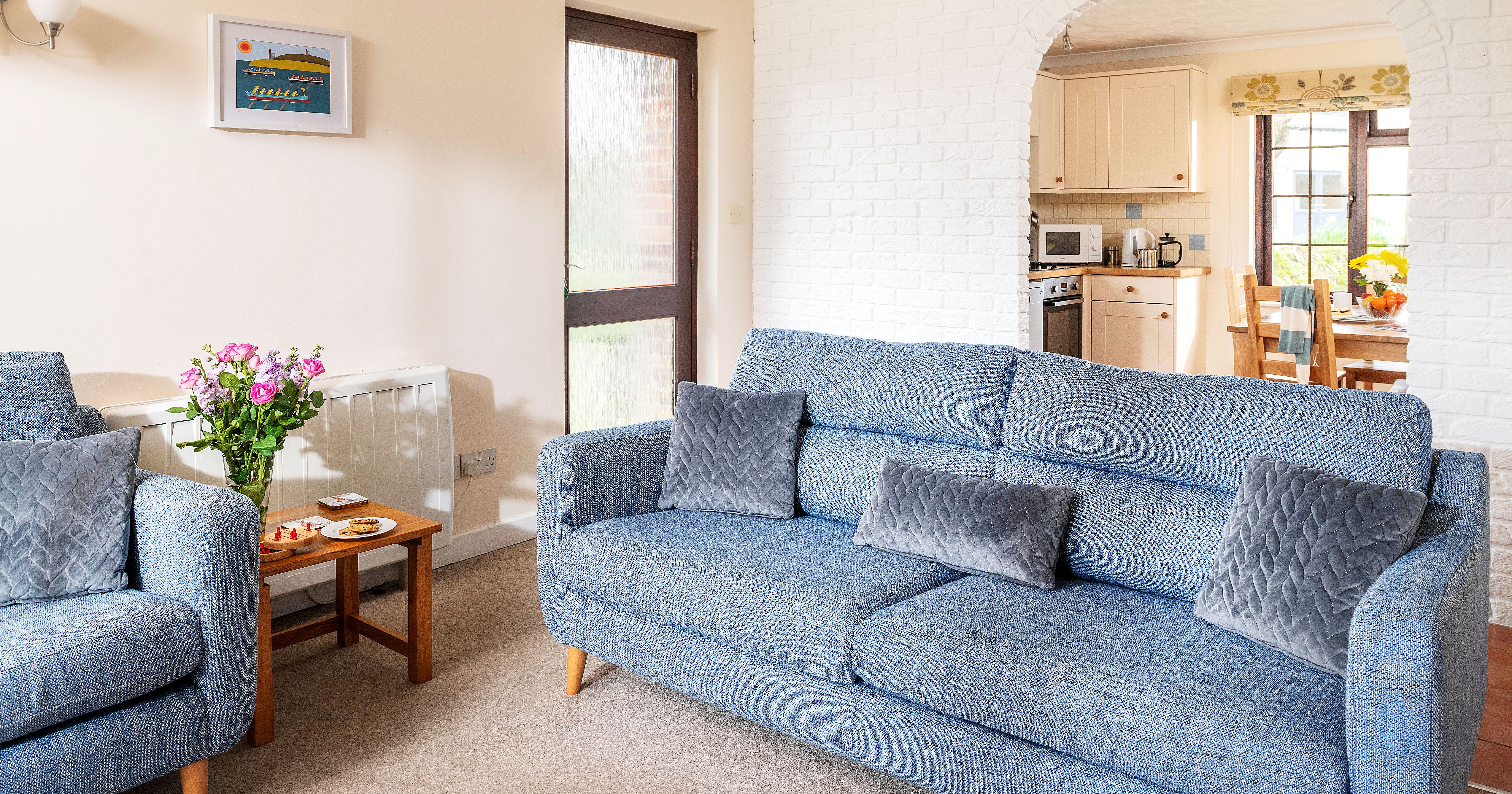 A cozy living room with blue sofas, gray quilted cushions, a wooden coffee table with flowers and snacks, and an arched doorway leading to a kitchen and dining area.
