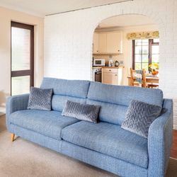 A cozy living room with blue sofas, gray quilted cushions, a wooden coffee table with flowers and snacks, and an arched doorway leading to a kitchen and dining area.