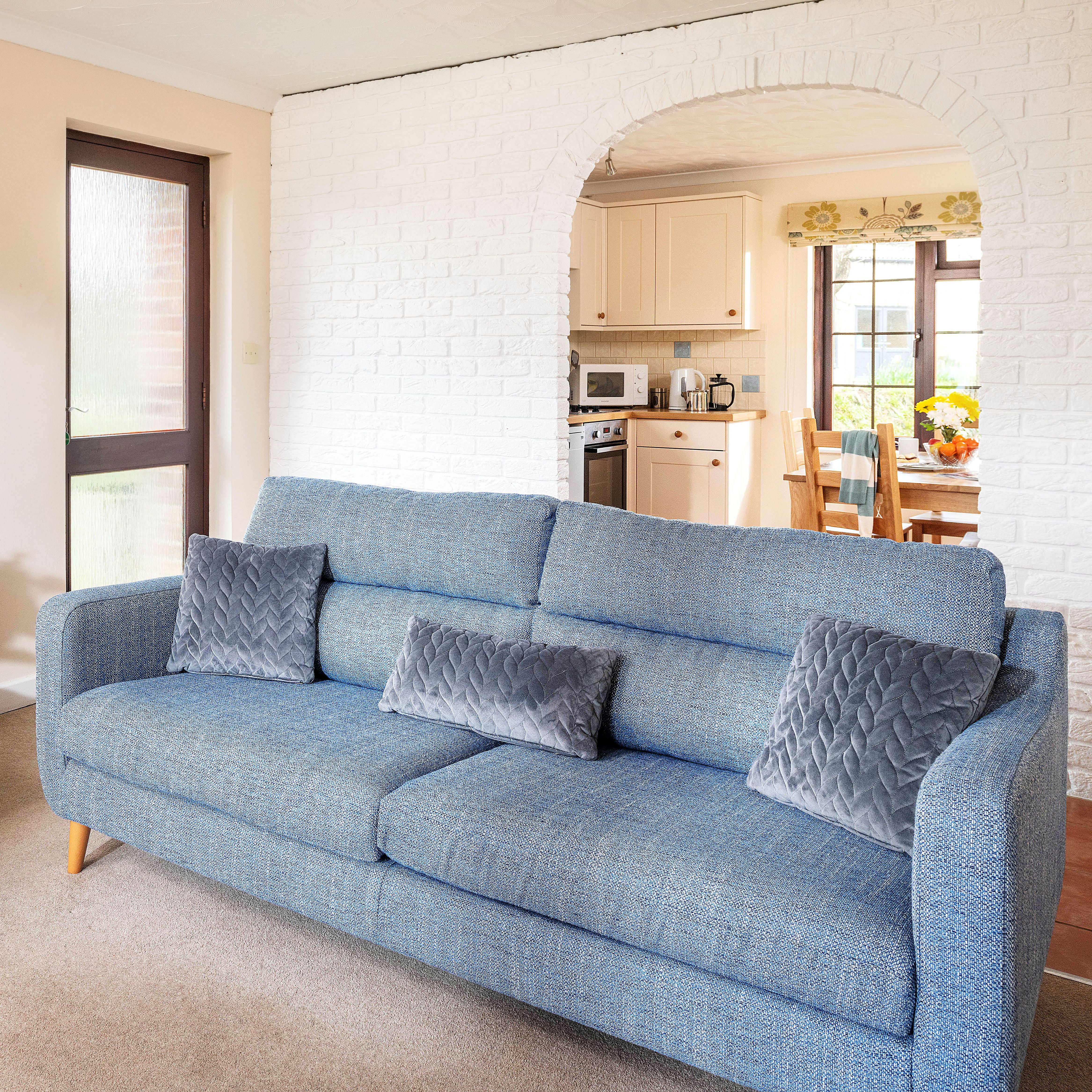 A cozy living room with blue sofas, gray quilted cushions, a wooden coffee table with flowers and snacks, and an arched doorway leading to a kitchen and dining area.