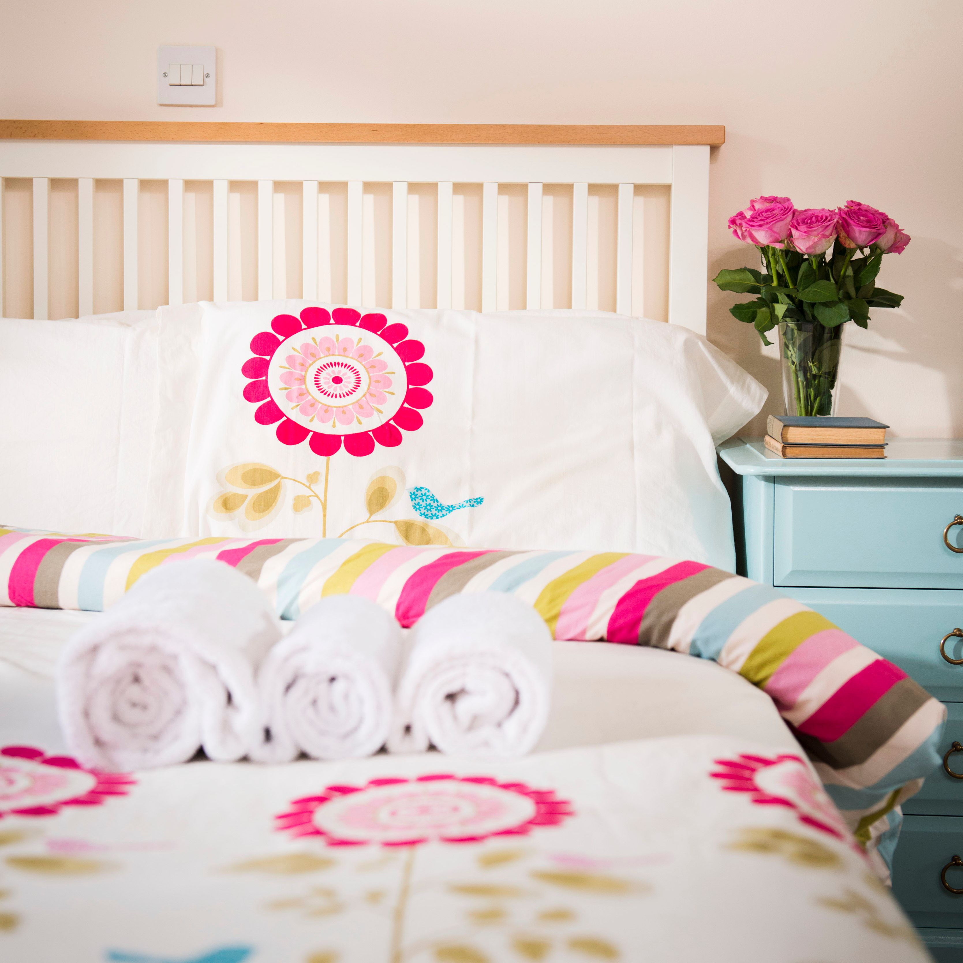 Neatly made bed with floral bedding, three rolled white towels, a blue nightstand with a vase of pink roses and stacked books, in a bright bedroom.