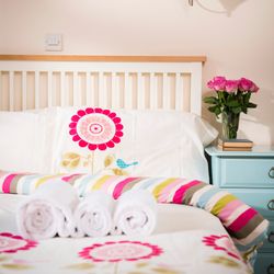 Neatly made bed with floral bedding, three rolled white towels, a blue nightstand with a vase of pink roses and stacked books, in a bright bedroom.
