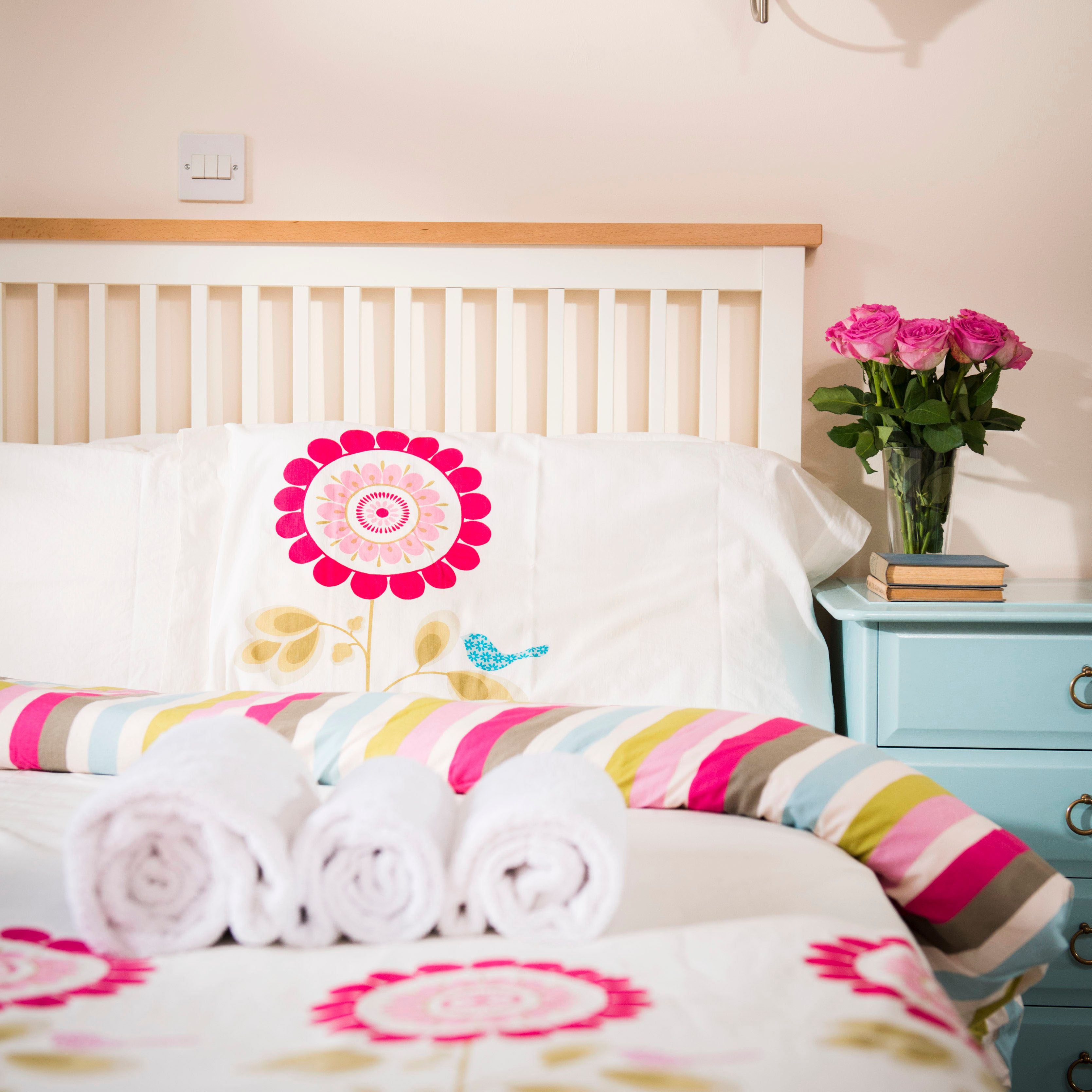 Neatly made bed with floral bedding, three rolled white towels, a blue nightstand with a vase of pink roses and stacked books, in a bright bedroom.