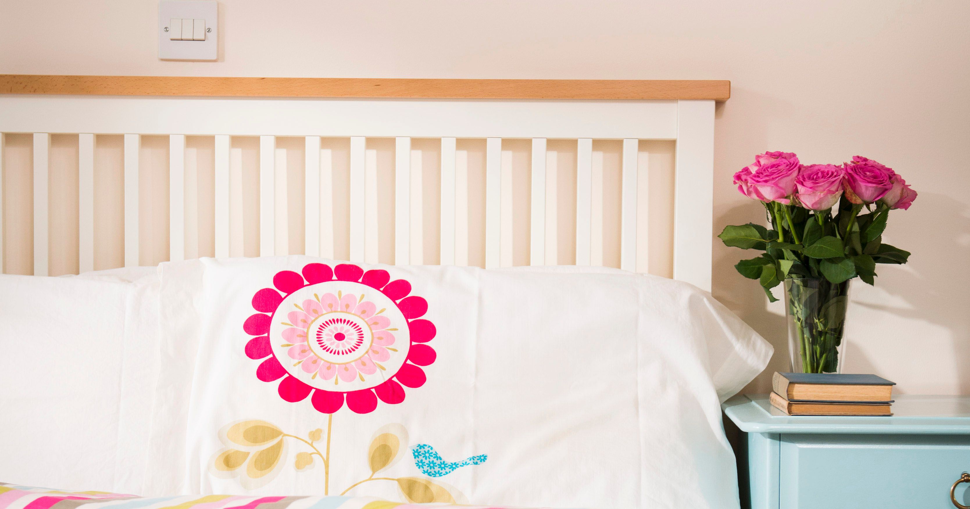 Neatly made bed with floral bedding, three rolled white towels, a blue nightstand with a vase of pink roses and stacked books, in a bright bedroom.