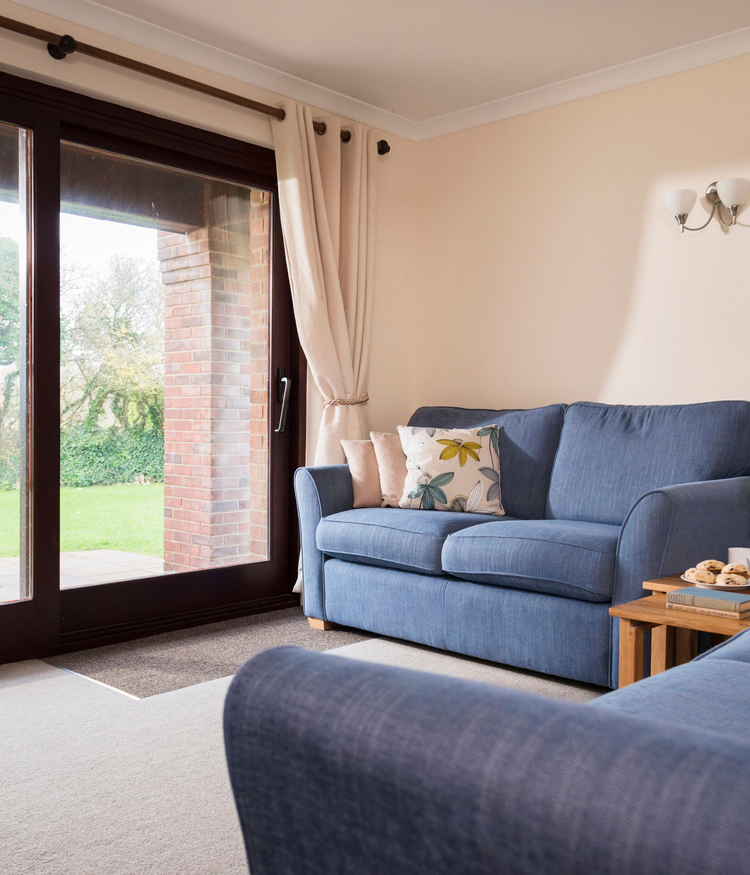 A cozy living room with two blue sofas, floral cushions, a wooden coffee table with flowers, mugs, and snacks, large sliding glass doors opening to a garden with a picnic table.
