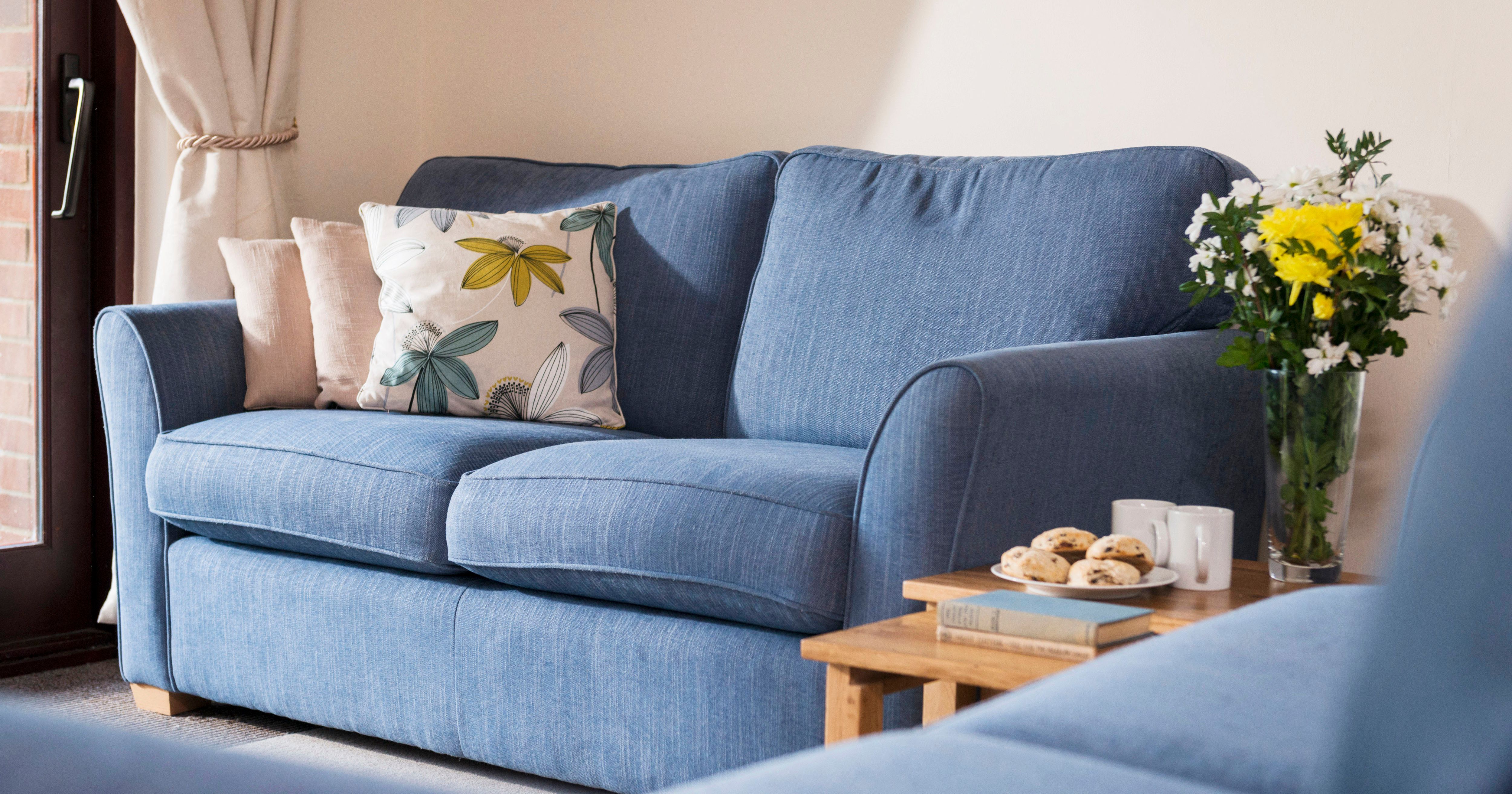 A cozy living room with a blue sofa, decorative pillows, a wooden side table with cookies, mugs, books, and a vase of fresh yellow and white flowers.