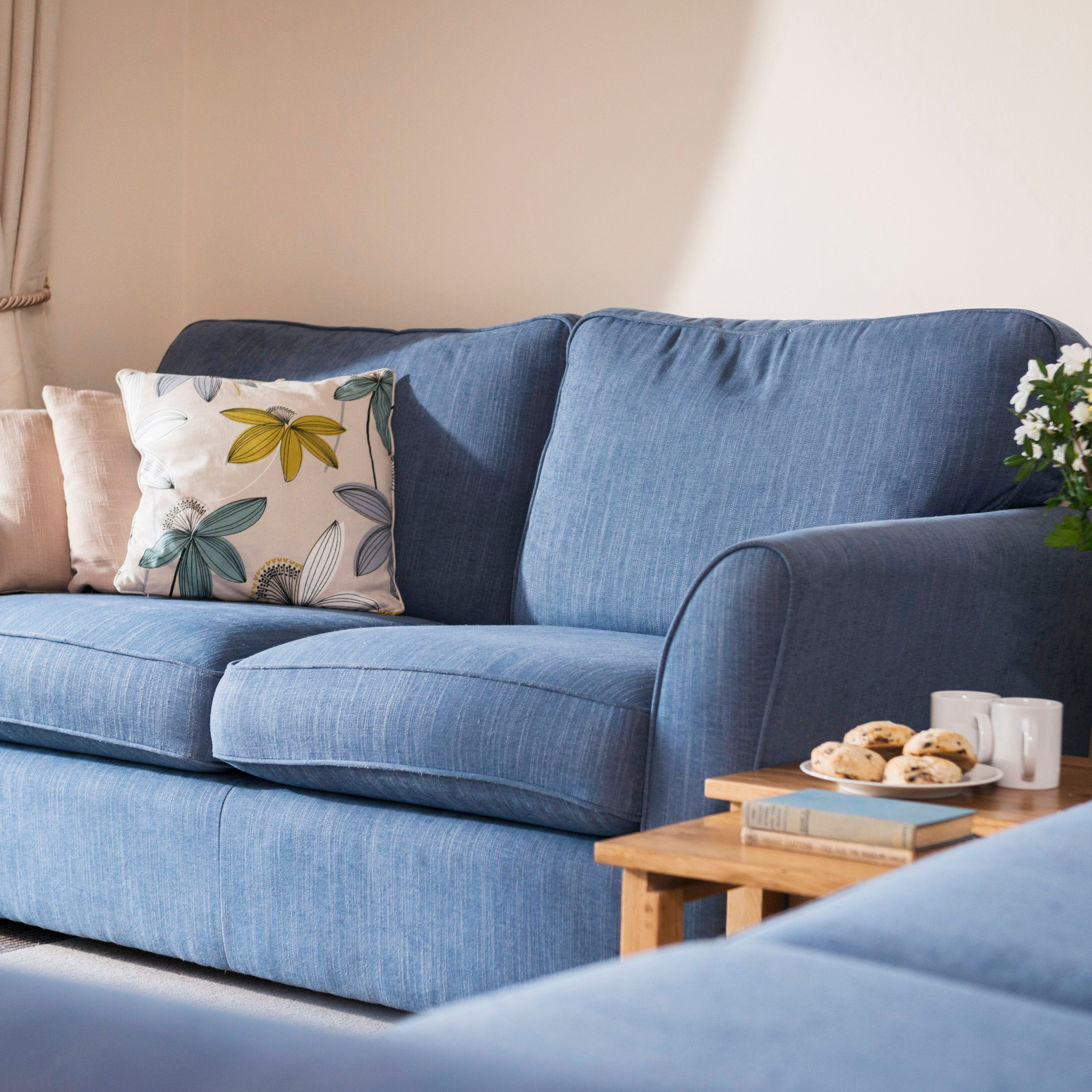 A cozy living room with a blue sofa, decorative pillows, a wooden side table with cookies, mugs, books, and a vase of fresh yellow and white flowers.