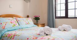 A cozy bedroom with a wooden bed frame, floral bedding, rolled white towels on the bed, a bedside table with a vase of pink flowers, and a window with blue curtains letting in natural light.