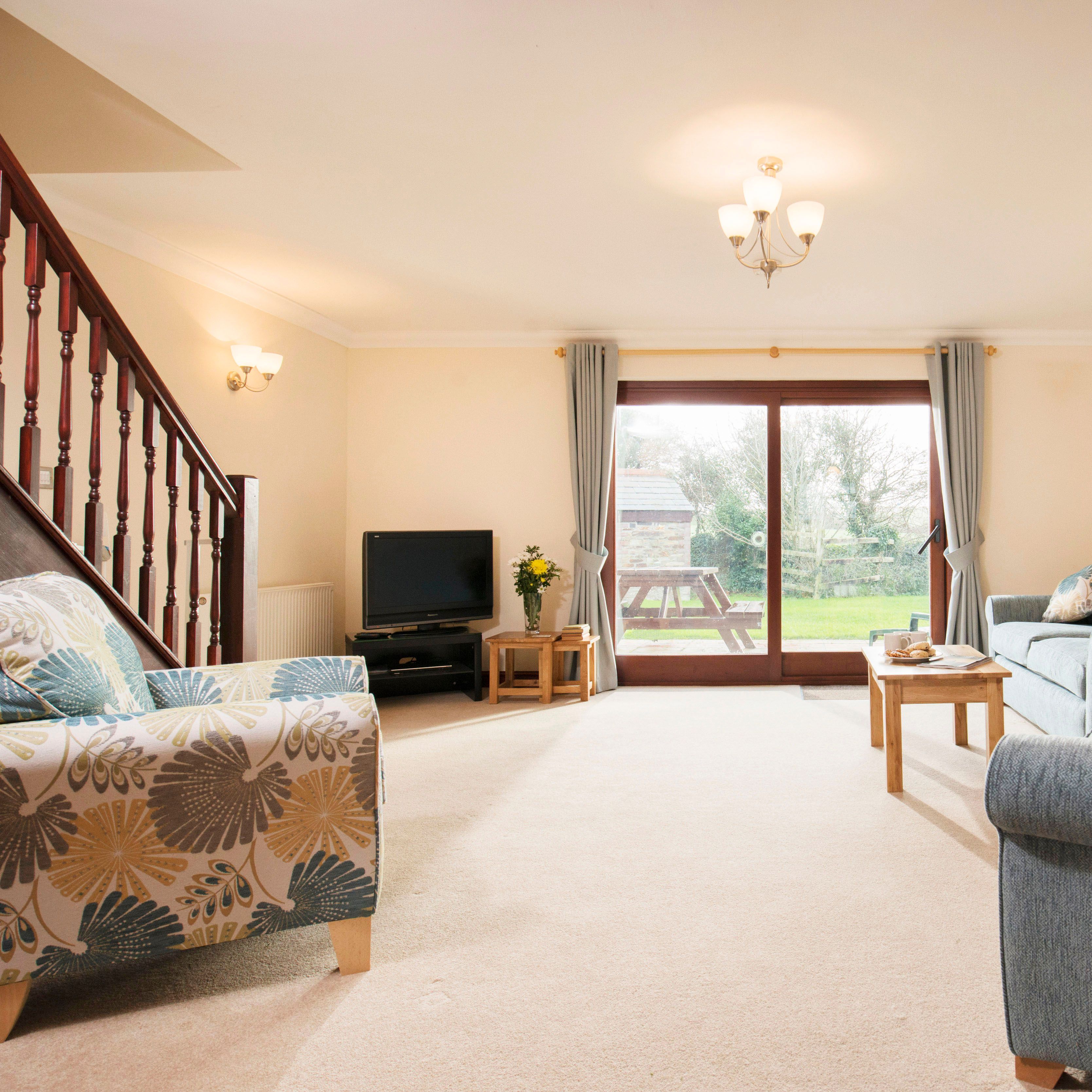 Bright and spacious living room with blue sofas, a patterned armchair, a wooden coffee table, TV, wooden staircase, and large sliding glass doors opening to a garden.