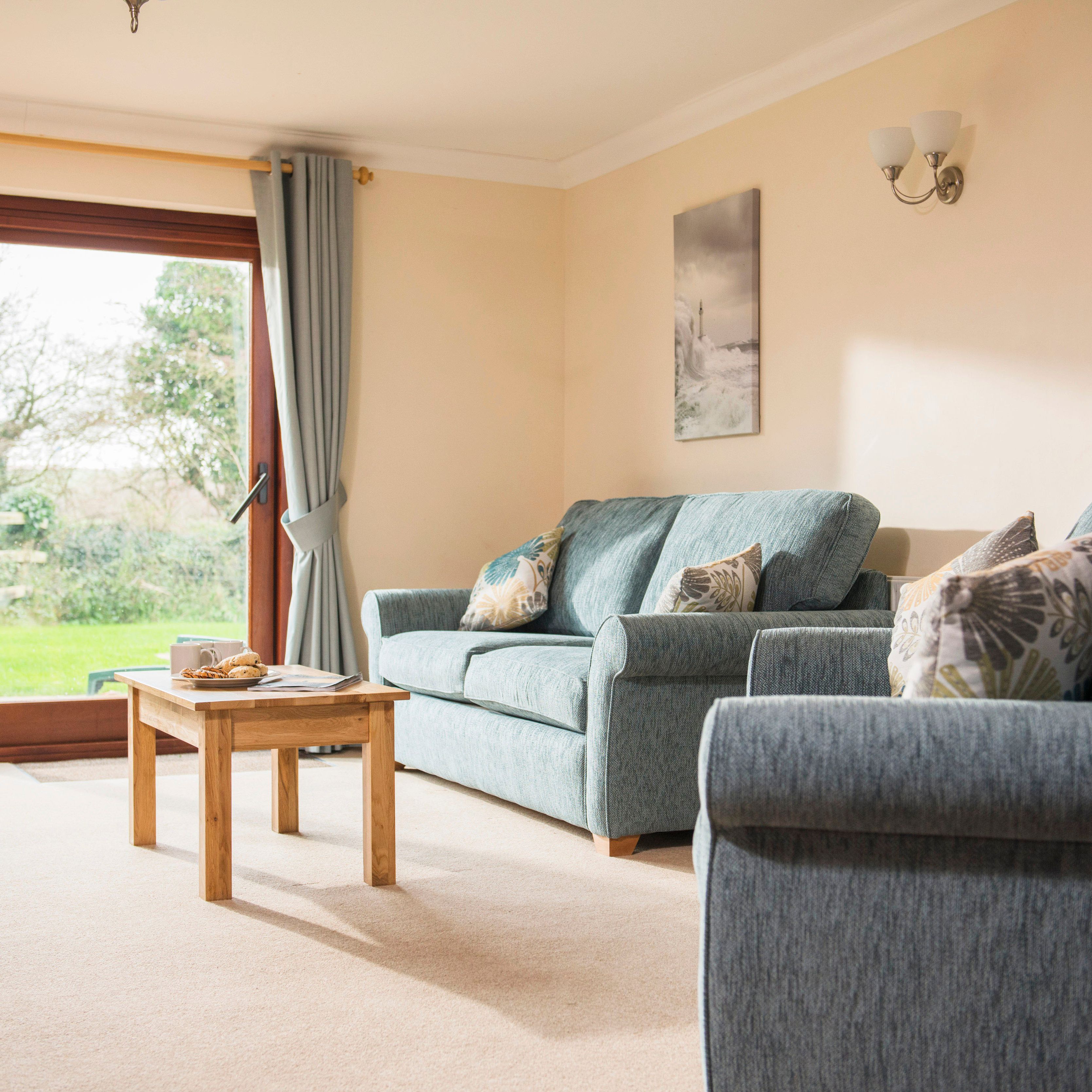 A bright and cozy living room with blue sofas, decorative pillows, a wooden coffee table, large glass doors with curtains, and photos on the wall. Outside the glass doors is a green garden.