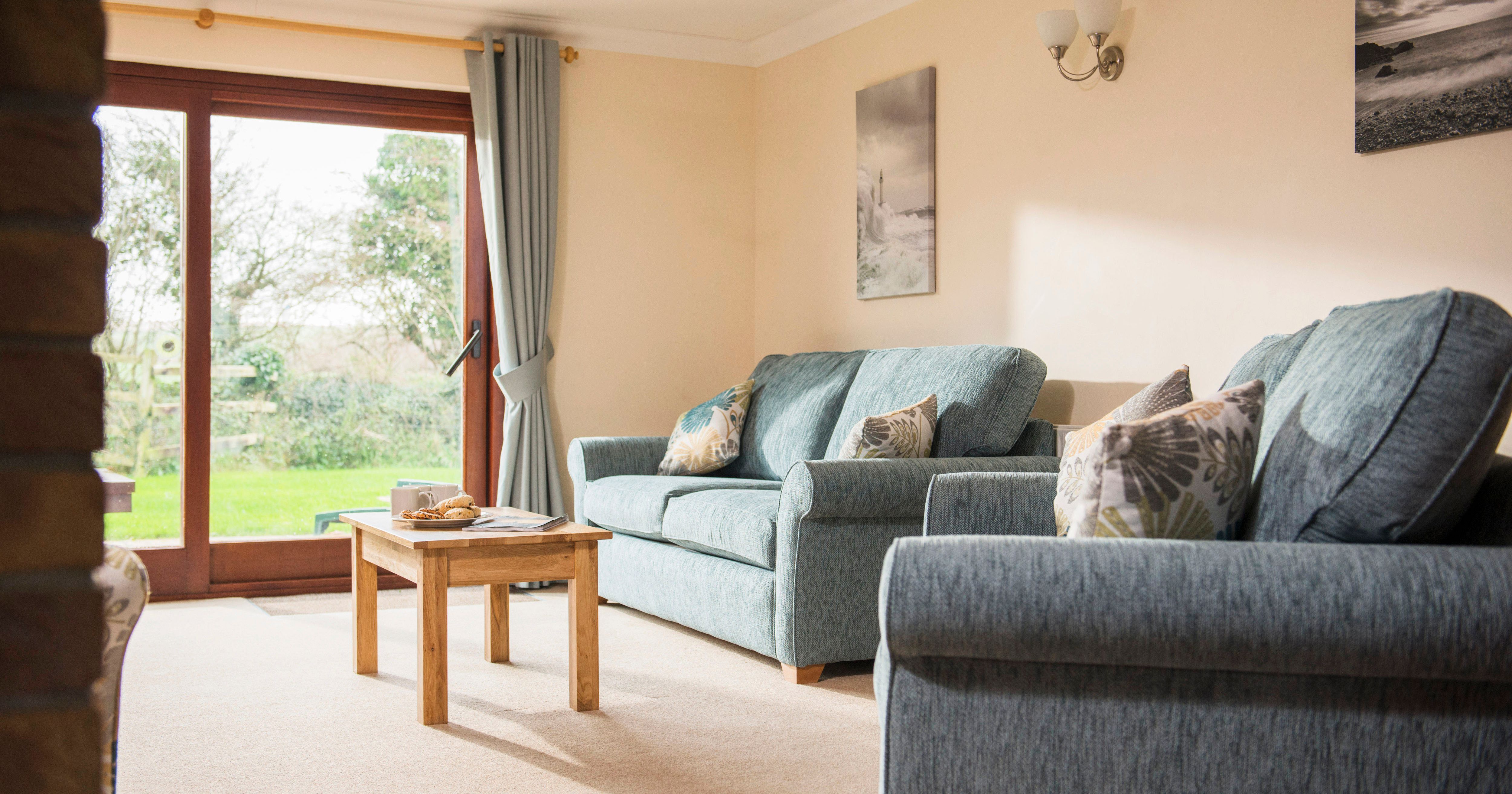 A bright and cozy living room with blue sofas, decorative pillows, a wooden coffee table, large glass doors with curtains, and photos on the wall. Outside the glass doors is a green garden.