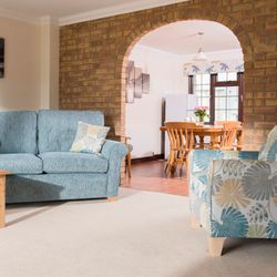 Cozy living room with a blue sofa, patterned armchair, wooden coffee table with snacks, and a dining area visible through a brick archway.