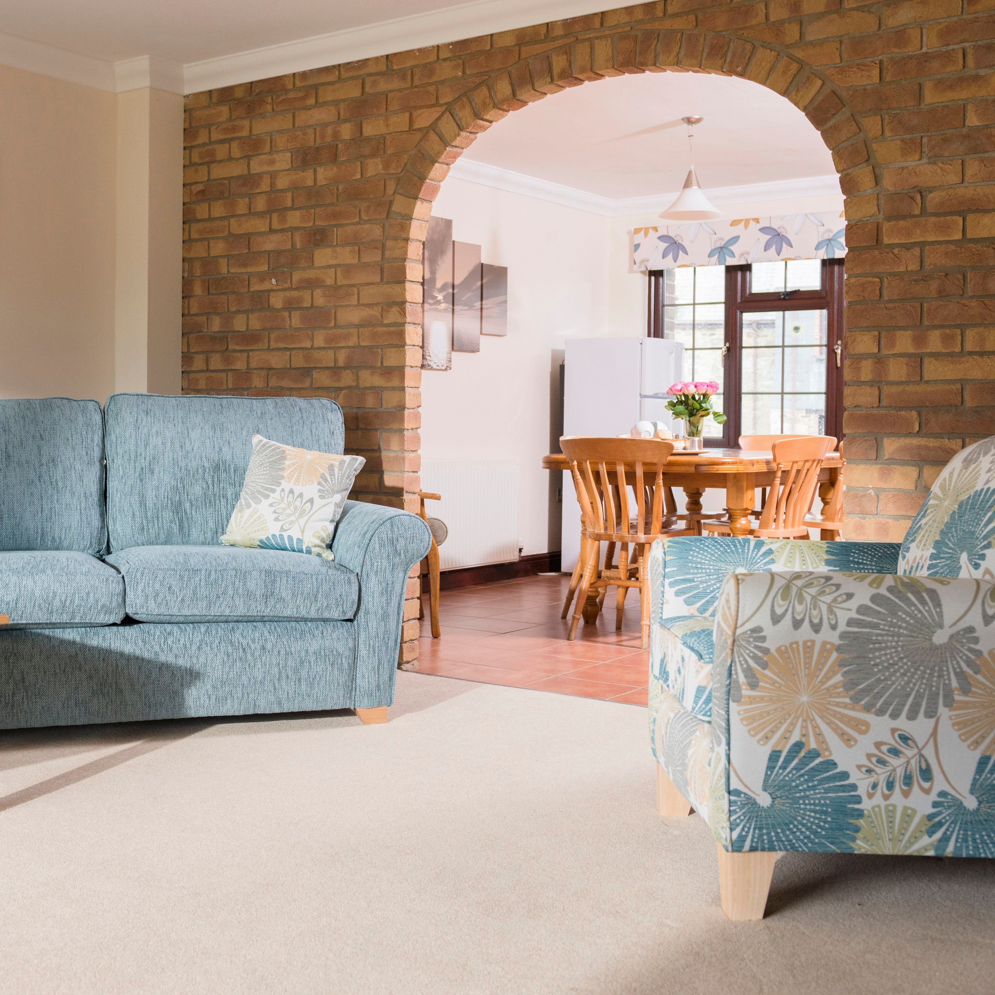Cozy living room with a blue sofa, patterned armchair, wooden coffee table with snacks, and a dining area visible through a brick archway.