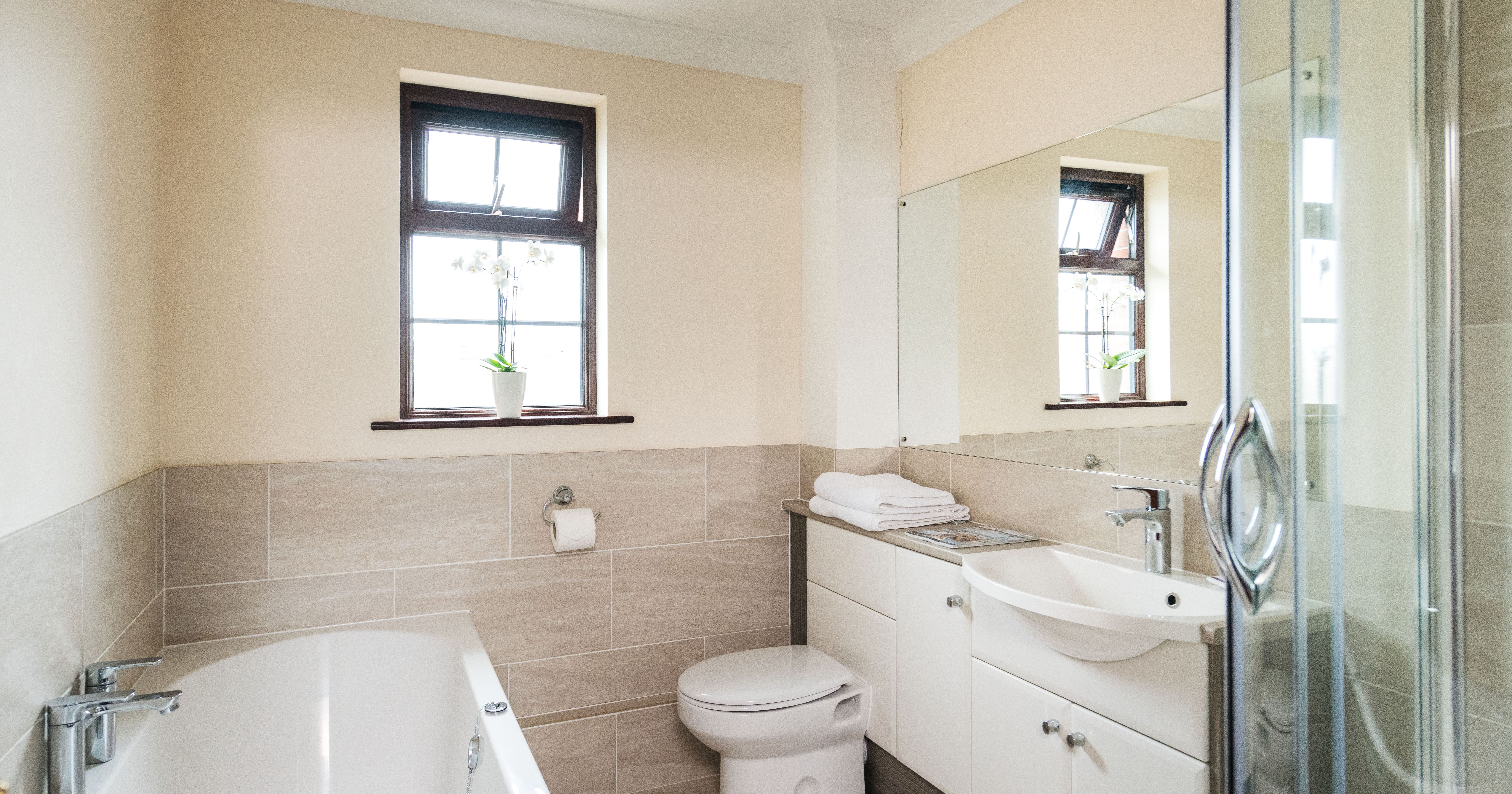 Modern bathroom with a bathtub, toilet, sink, glass shower enclosure, large mirror, and two windows with potted plants.