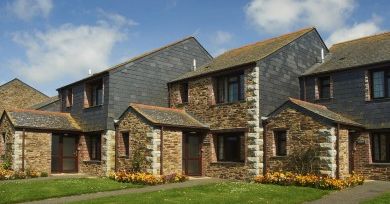 Row of modern stone houses with slate roofs, front lawns, and flower beds under a blue sky.