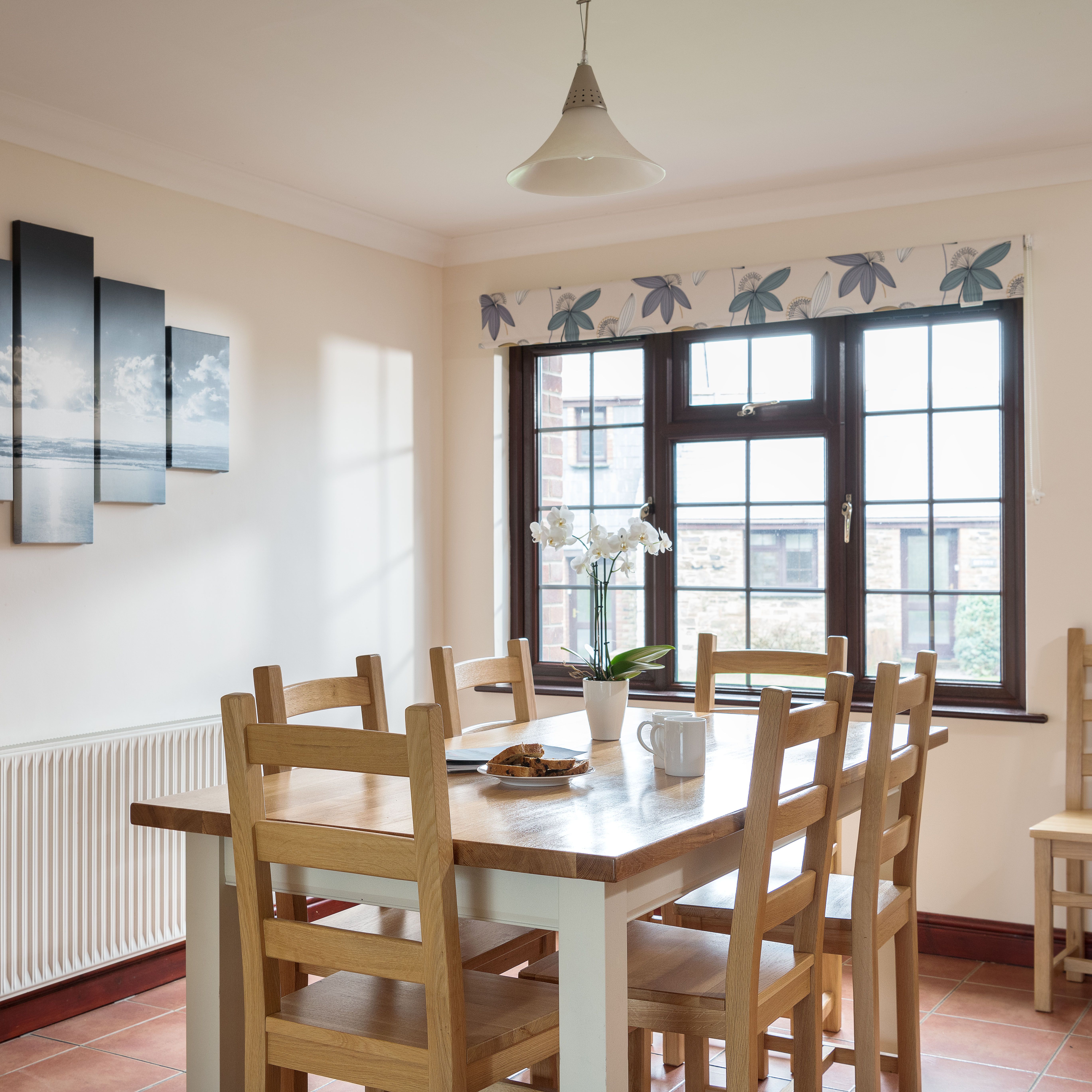 Bright dining room with wooden table and chairs, a window with floral valance, a modern pendant light, wall art depicting a seascape, and a white orchid in a vase on the table.