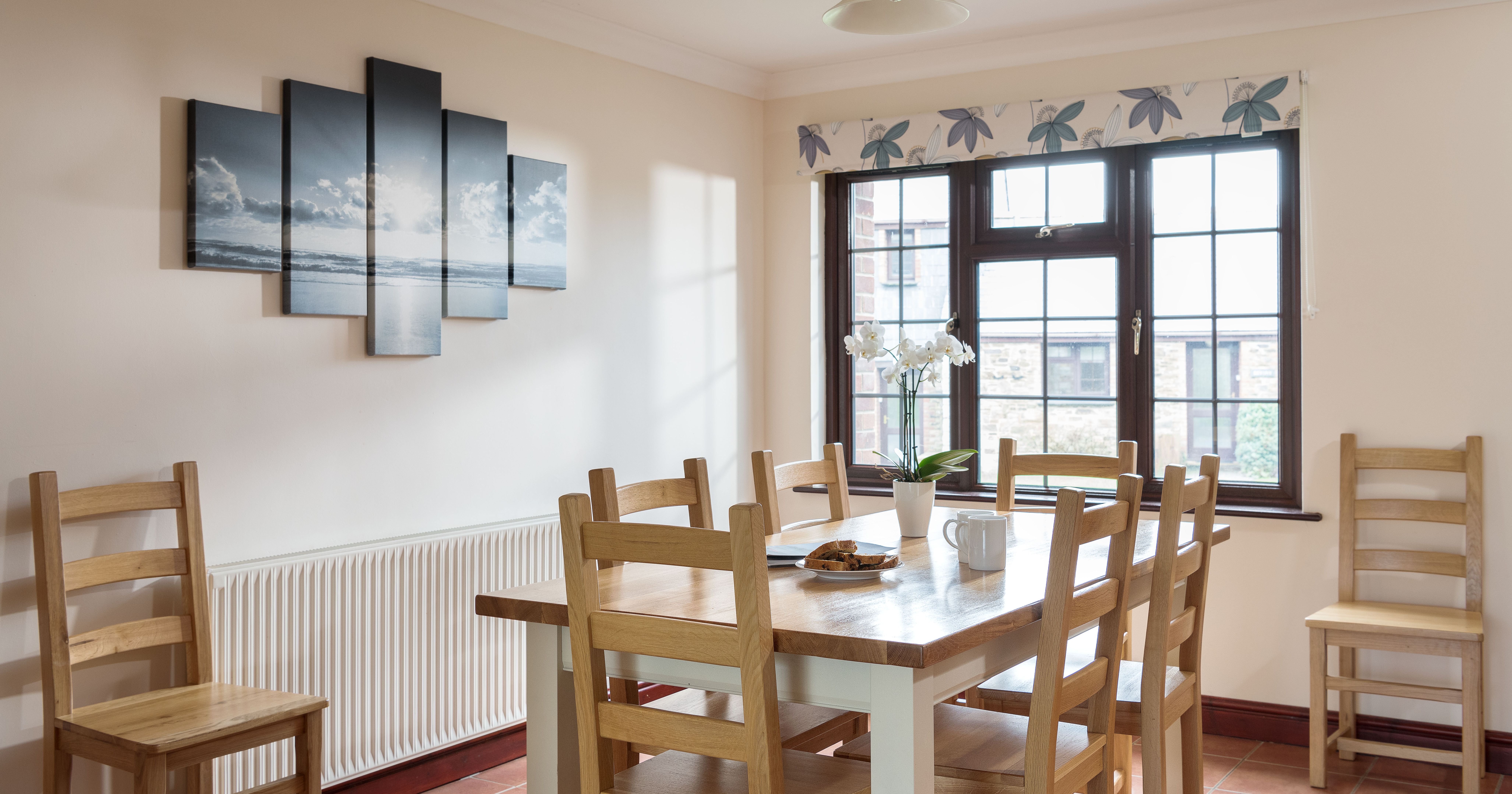 Bright dining room with wooden table and chairs, a window with floral valance, a modern pendant light, wall art depicting a seascape, and a white orchid in a vase on the table.