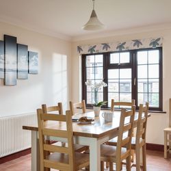 Bright dining room with wooden table and chairs, a window with floral valance, a modern pendant light, wall art depicting a seascape, and a white orchid in a vase on the table.