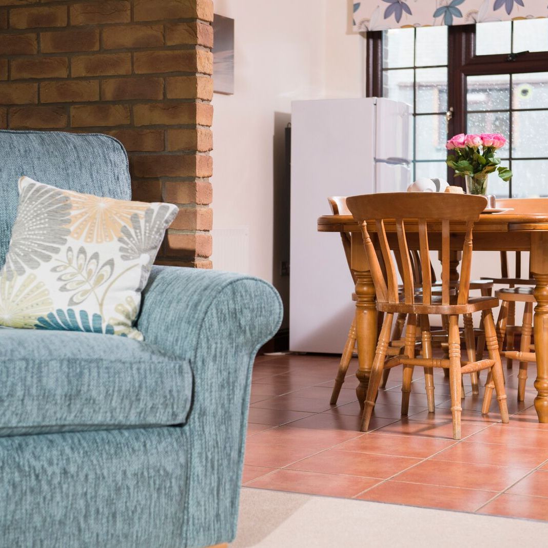 Cozy living and dining area with a blue sofa, patterned cushion, wooden dining table with six chairs, refrigerator, and a vase of pink flowers on the table near a large window.