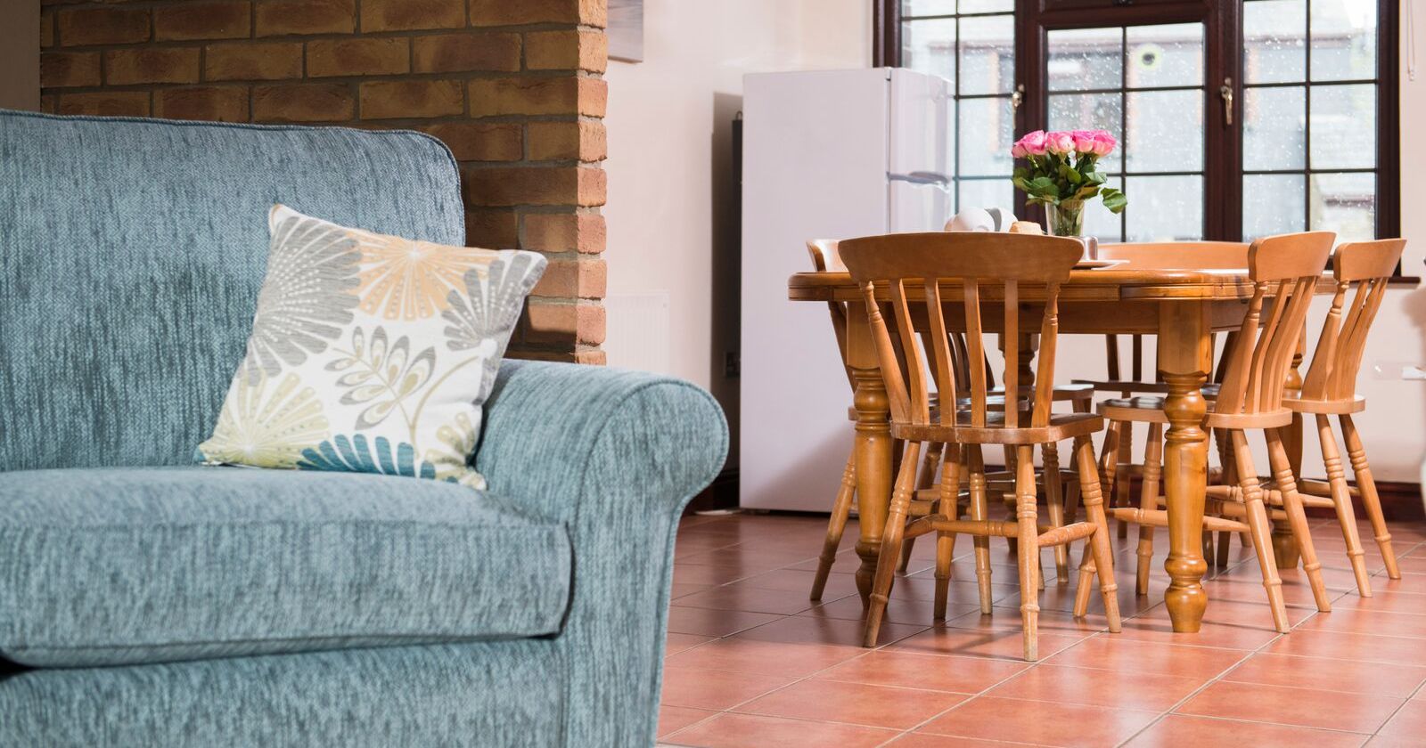 Cozy living and dining area with a blue sofa, patterned cushion, wooden dining table with six chairs, refrigerator, and a vase of pink flowers on the table near a large window.