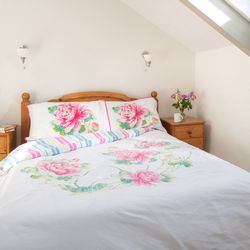 Bright bedroom with a wooden bed, floral bedding, two wooden nightstands, a vase of flowers, books, and a window with light curtains.