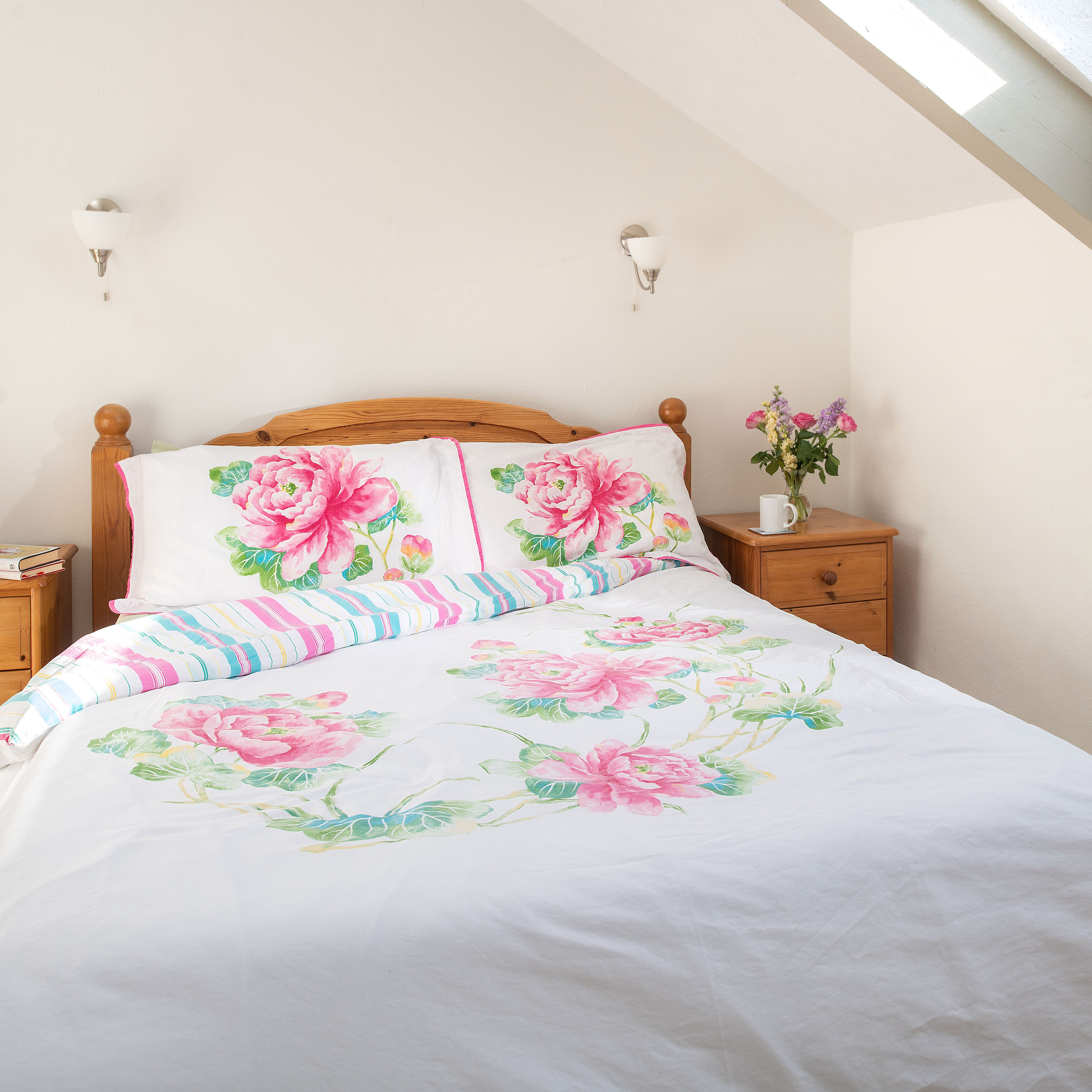 Bright bedroom with a wooden bed, floral bedding, two wooden nightstands, a vase of flowers, books, and a window with light curtains.
