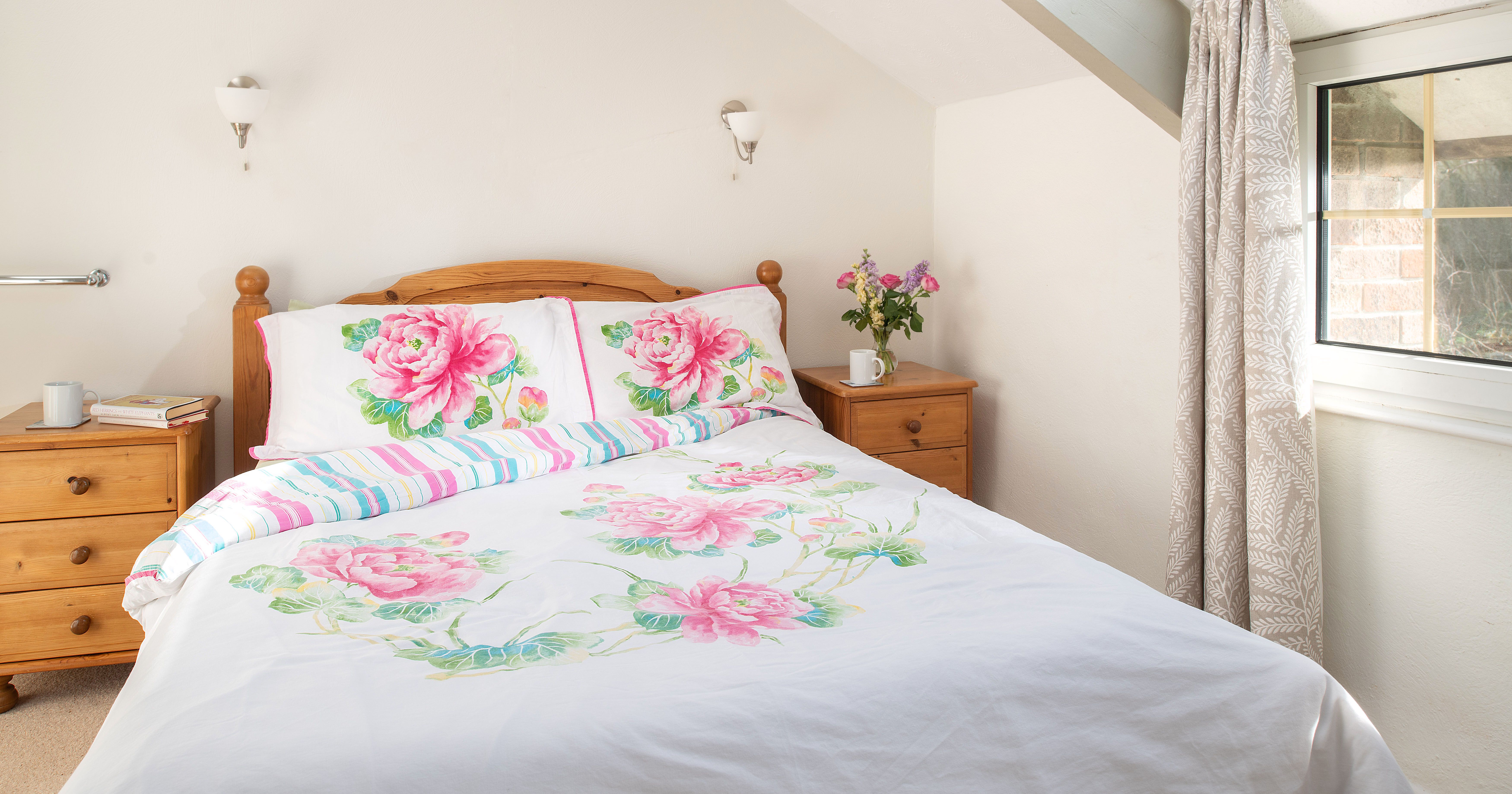 Bright bedroom with a wooden bed, floral bedding, two wooden nightstands, a vase of flowers, books, and a window with light curtains.