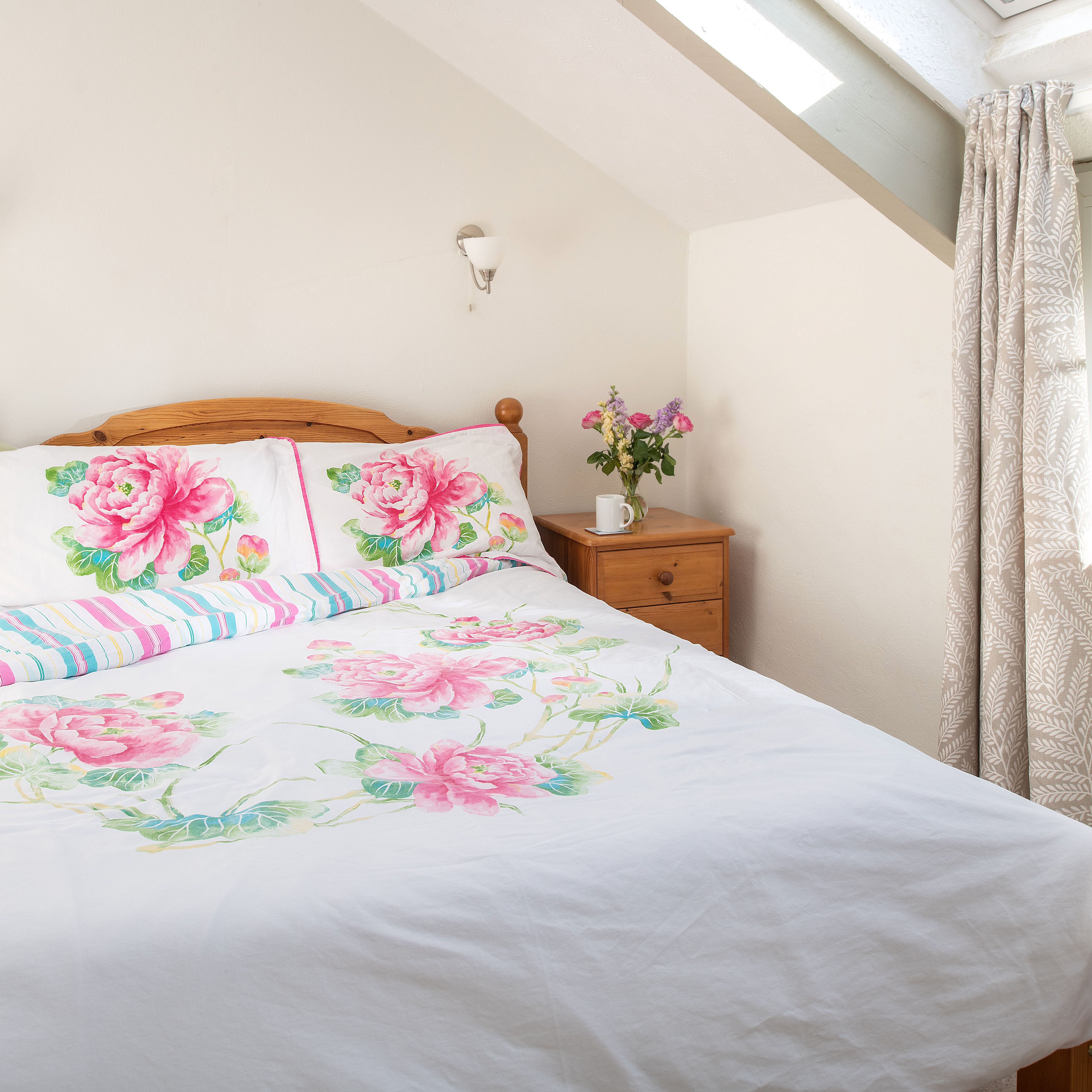 Bright bedroom with a wooden bed, floral bedding, two wooden nightstands, a vase of flowers, books, and a window with light curtains.