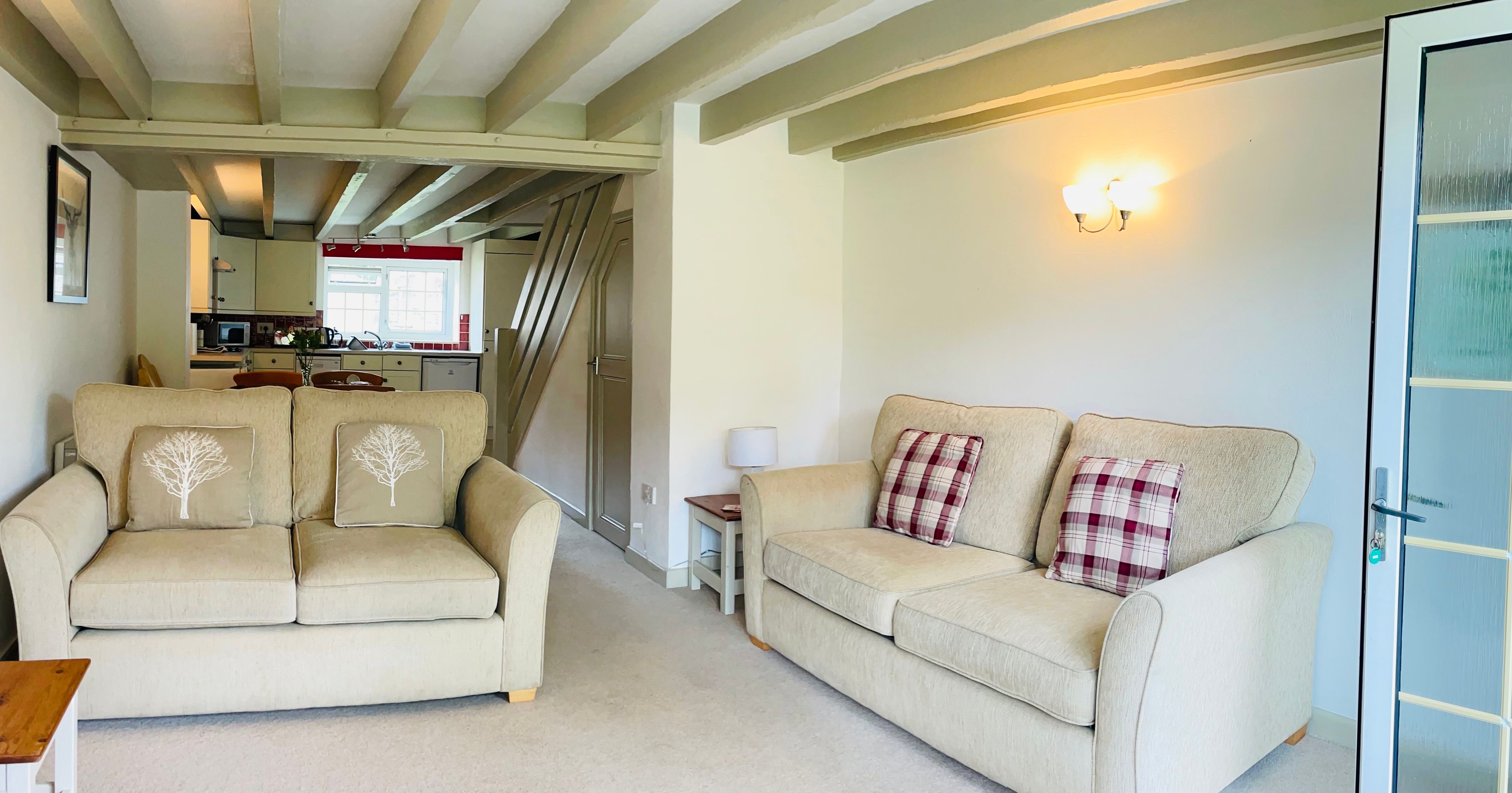 Cozy living room with two beige sofas, decorative cushions, wooden beams on the ceiling, wall-mounted light fixture, and a view into a kitchen and dining area in the background.