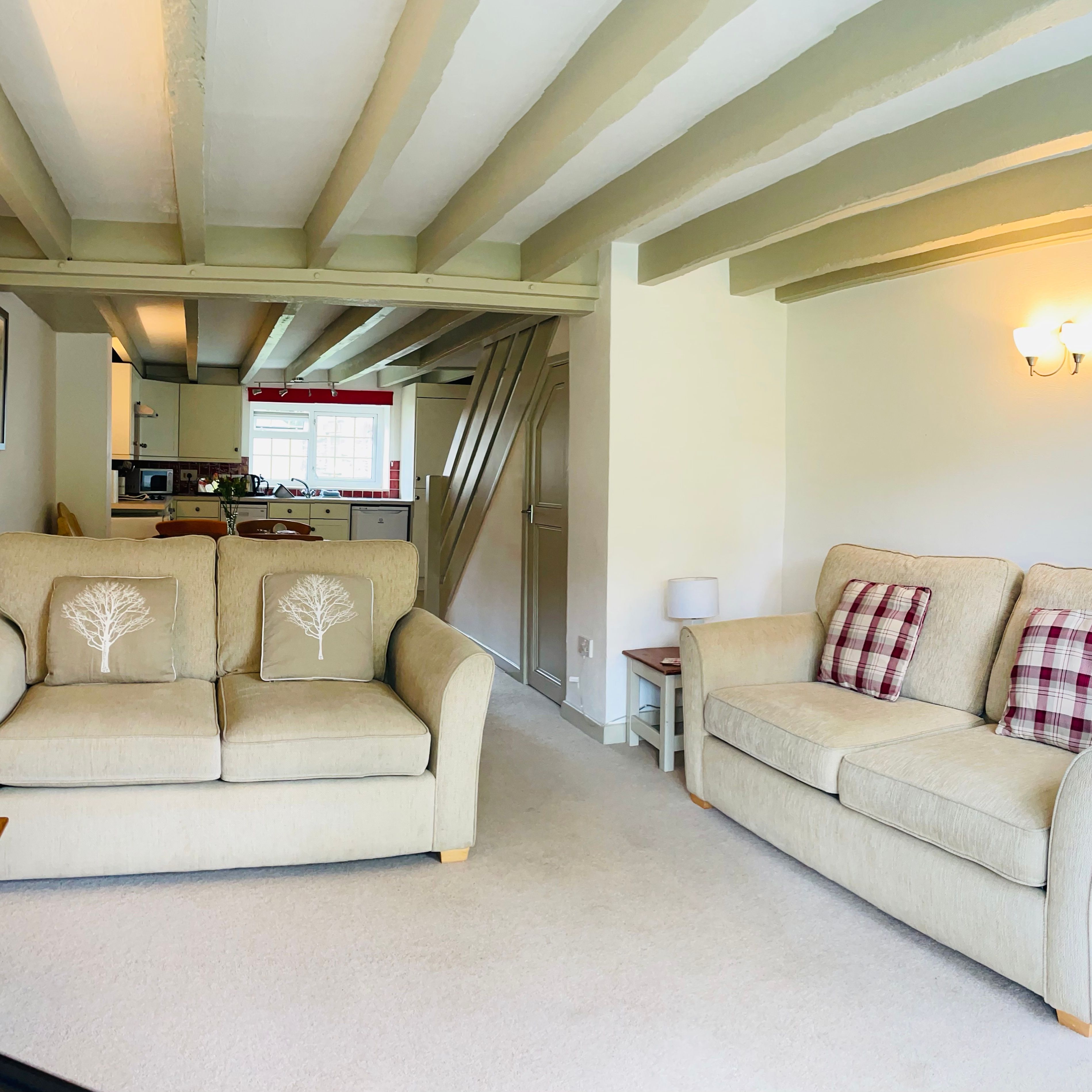 Cozy living room with two beige sofas, decorative cushions, wooden beams on the ceiling, wall-mounted light fixture, and a view into a kitchen and dining area in the background.