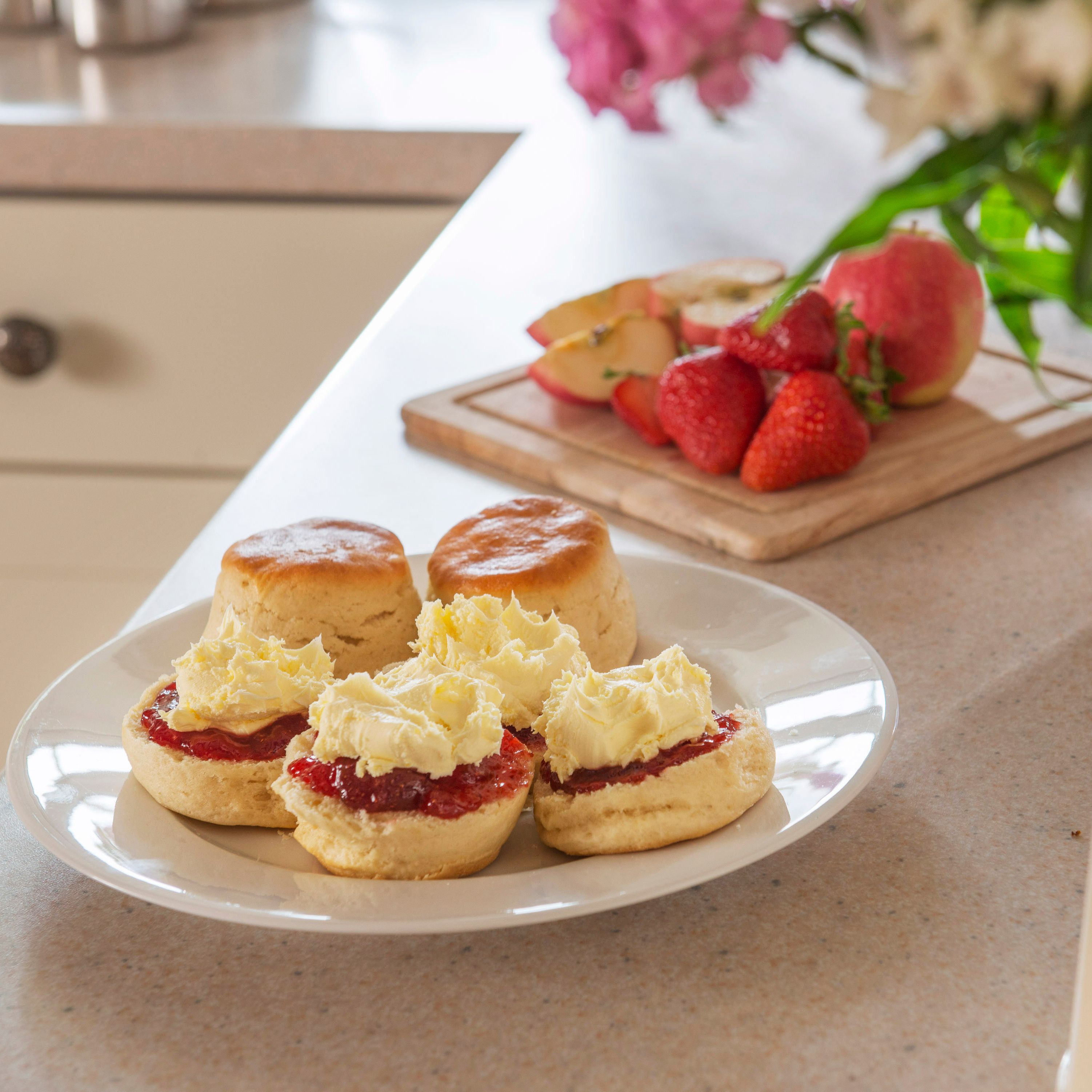 A plate of scones with clotted cream and jam on a kitchen counter, with fresh strawberries and sliced apples on a wooden board in the background, and a jug of flowers in the foreground.