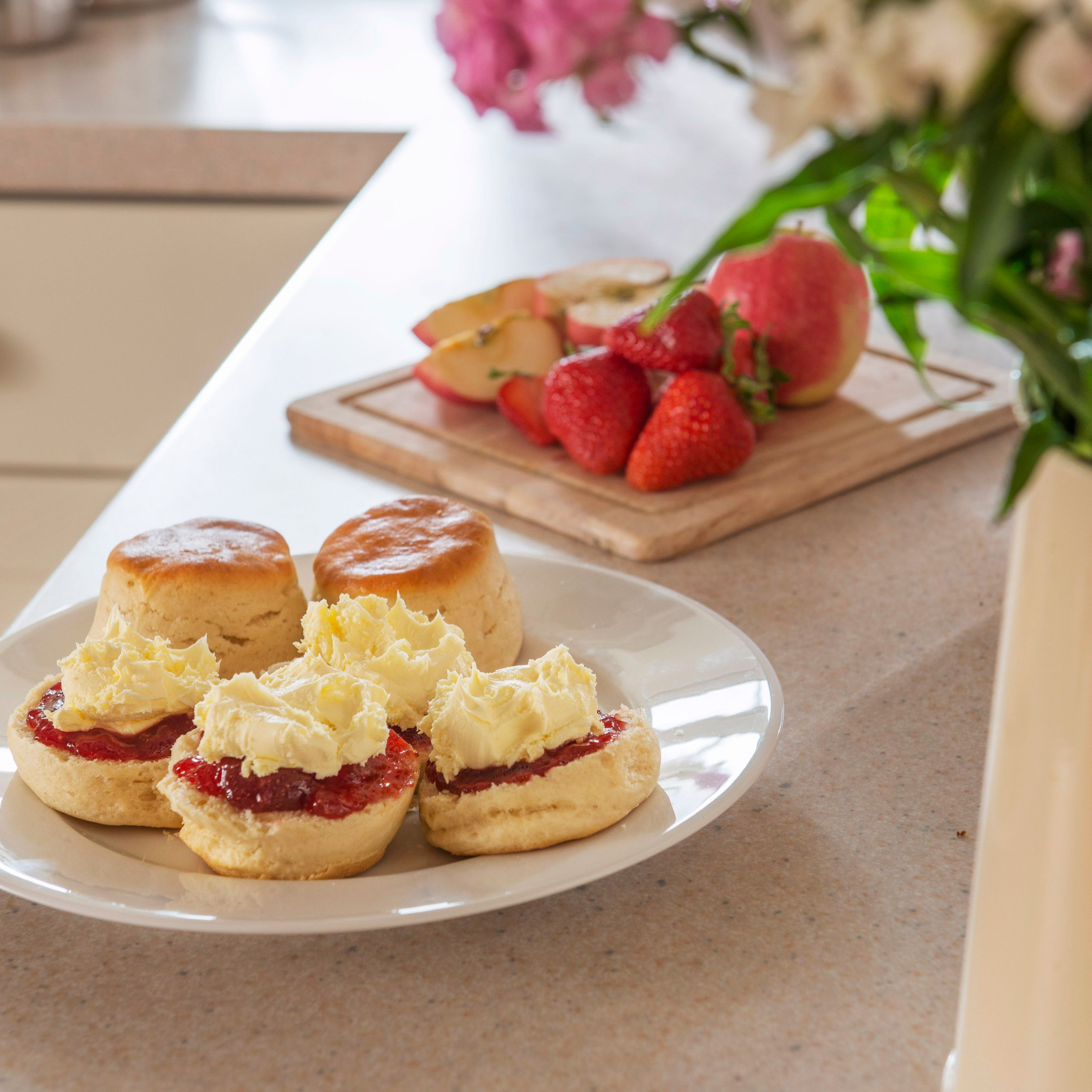 A plate of scones with clotted cream and jam on a kitchen counter, with fresh strawberries and sliced apples on a wooden board in the background, and a jug of flowers in the foreground.