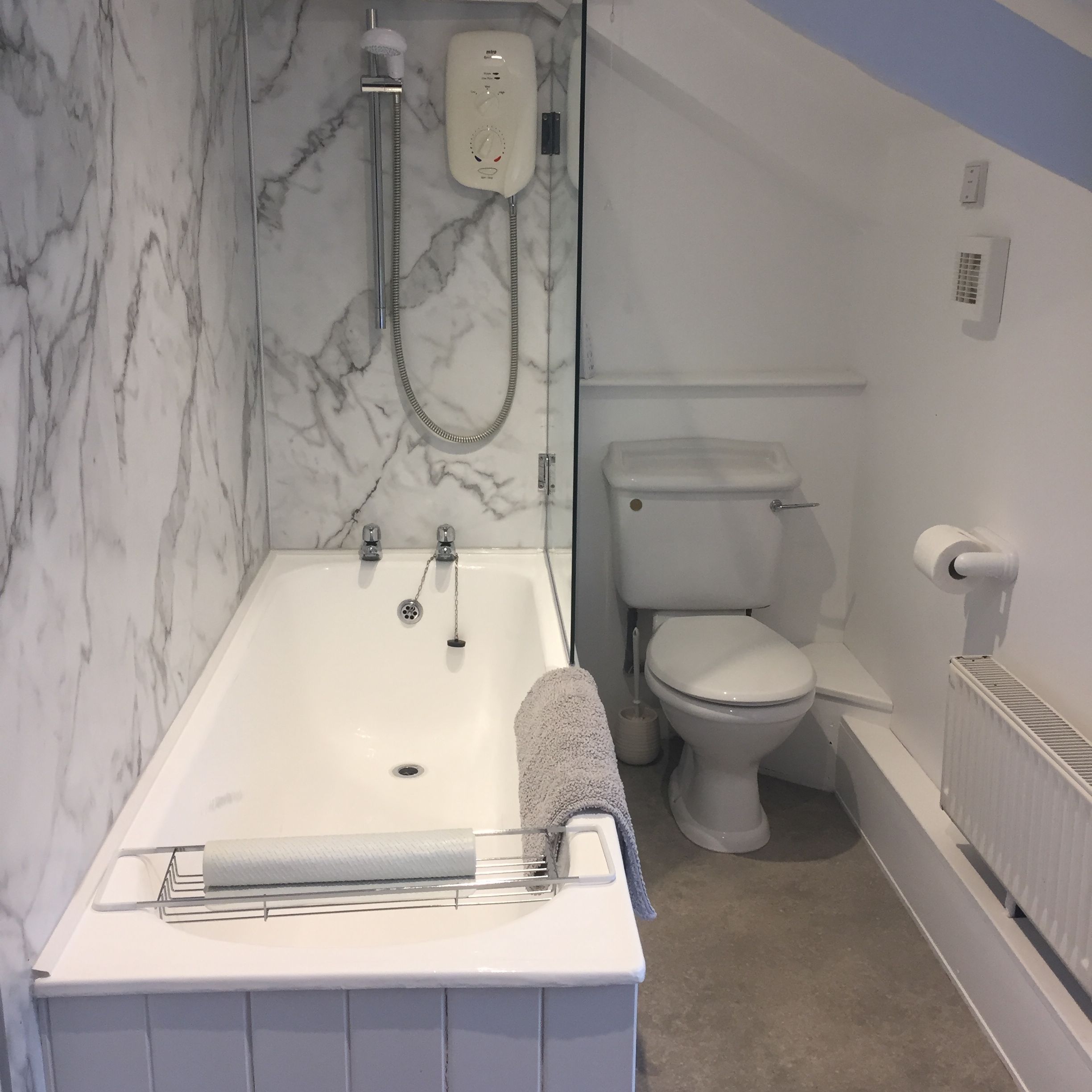 A modern bathroom with a white bathtub, marble-patterned wall, shower with electric unit, toilet, towel on the edge of the tub, toilet paper holder, radiator, and sloped ceiling.