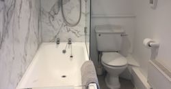A modern bathroom with a white bathtub, marble-patterned wall, shower with electric unit, toilet, towel on the edge of the tub, toilet paper holder, radiator, and sloped ceiling.