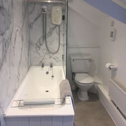 A modern bathroom with a white bathtub, marble-patterned wall, shower with electric unit, toilet, towel on the edge of the tub, toilet paper holder, radiator, and sloped ceiling.