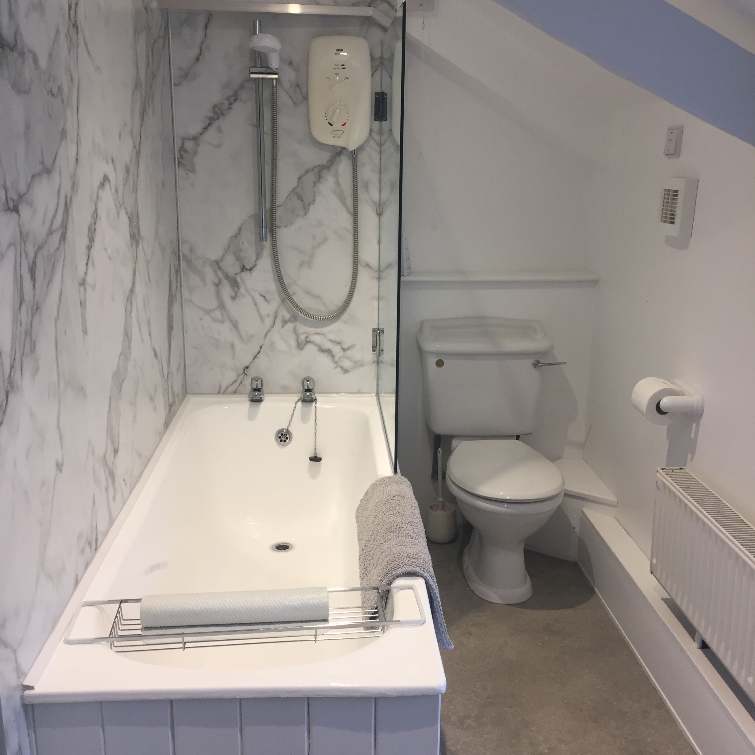 A modern bathroom with a white bathtub, marble-patterned wall, shower with electric unit, toilet, towel on the edge of the tub, toilet paper holder, radiator, and sloped ceiling.