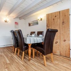 Dining room with wooden floor, round table covered with a gray polka dot tablecloth, four dark brown leather chairs, rustic wooden door, white walls, and artwork on the walls.