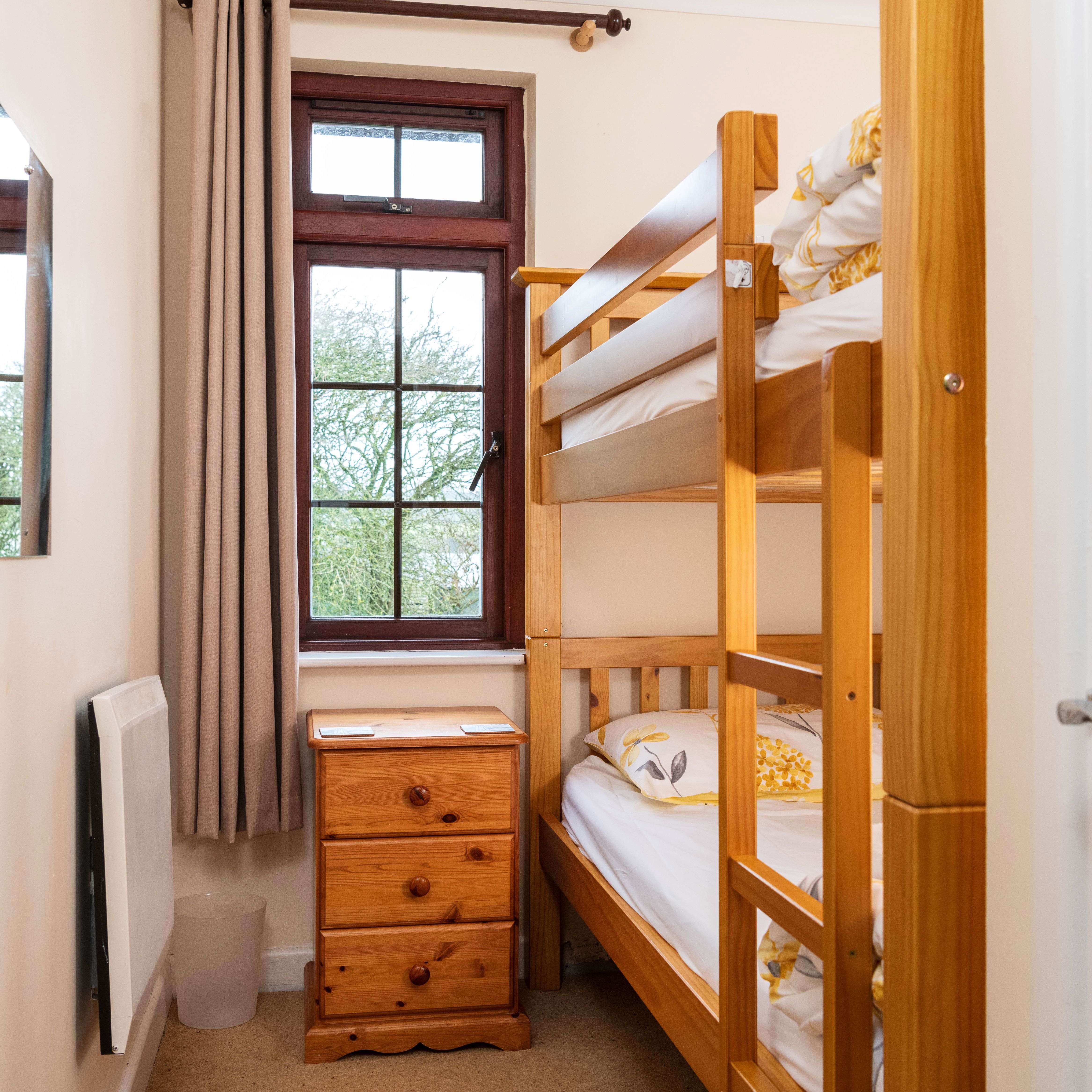 A small bedroom with a wooden bunk bed, a wooden nightstand with drawers, a window with brown curtains, a wall mirror, and a white heater.