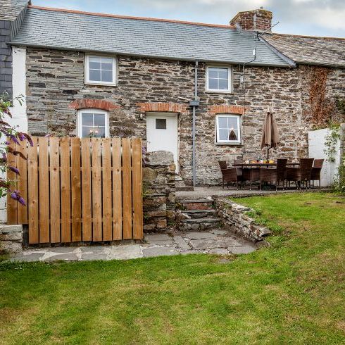 Traditional stone cottage with a slate roof, a small wooden gate, outdoor dining area with chairs and an umbrella, and a green lawn in front.