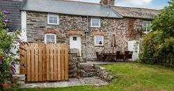 Traditional stone cottage with a slate roof, a small wooden gate, outdoor dining area with chairs and an umbrella, and a green lawn in front.