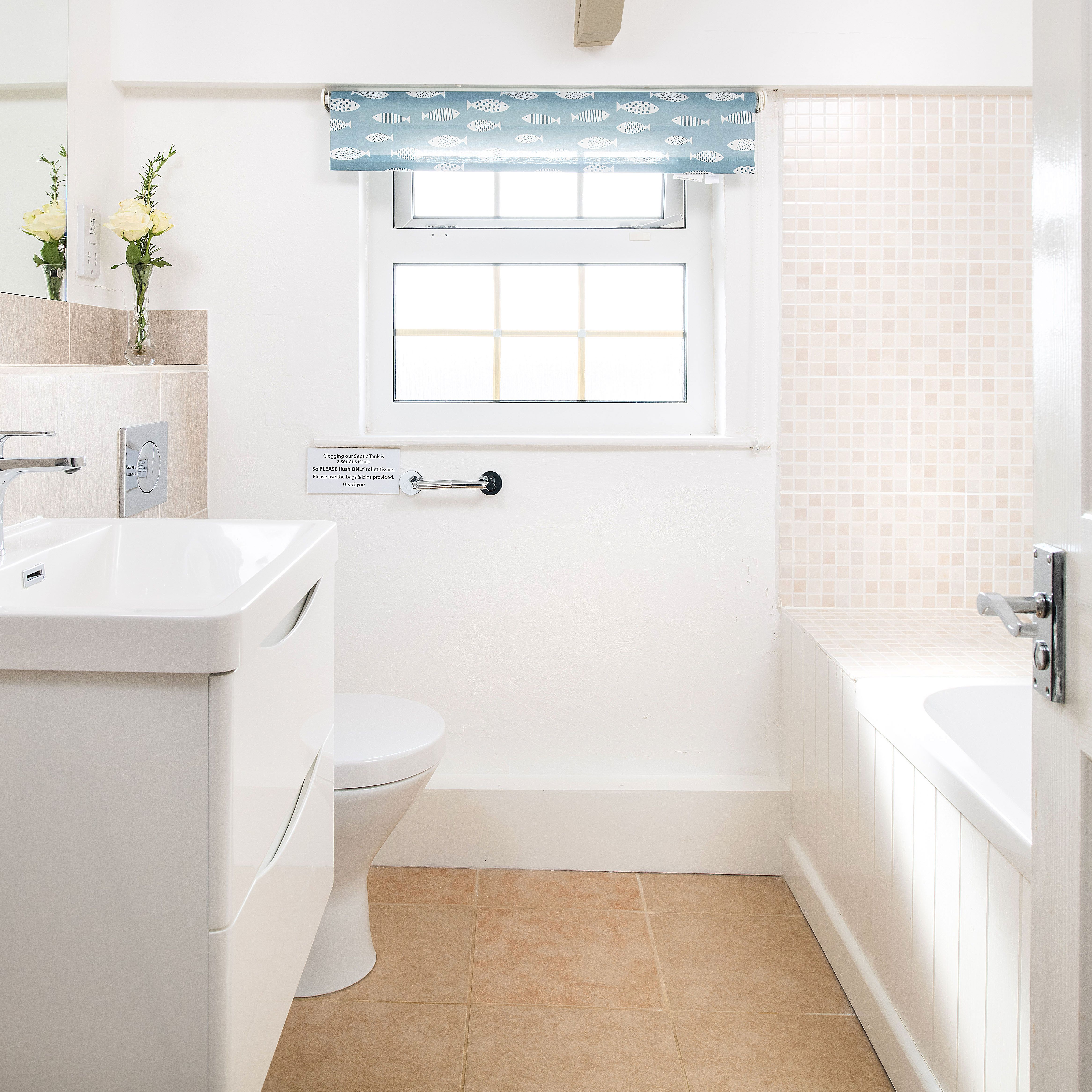 Bright, modern bathroom with white sink, toilet, and bathtub, beige tiles, a window with a blue patterned blind, and a vase with yellow flowers on the sink counter.