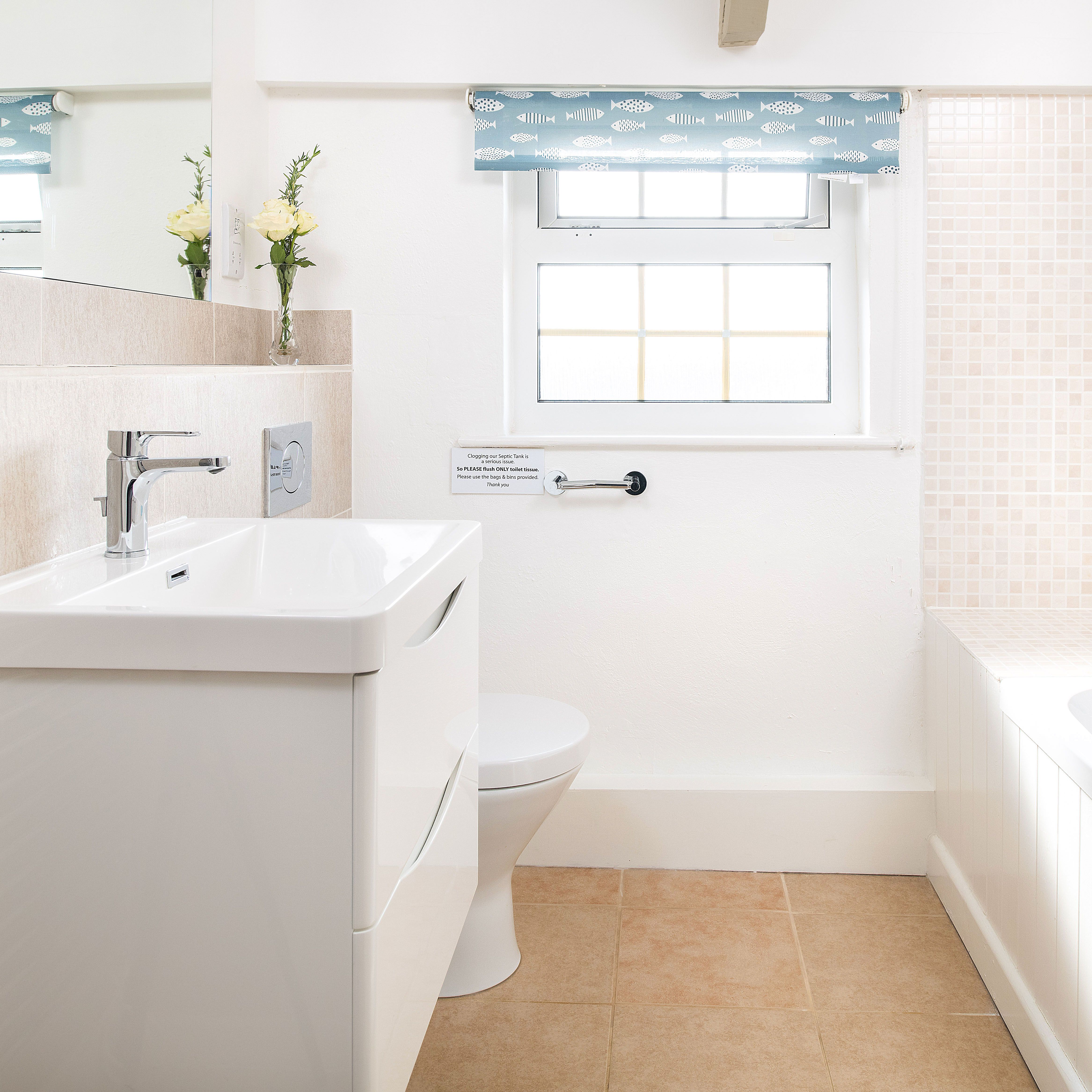 Bright, modern bathroom with white sink, toilet, and bathtub, beige tiles, a window with a blue patterned blind, and a vase with yellow flowers on the sink counter.