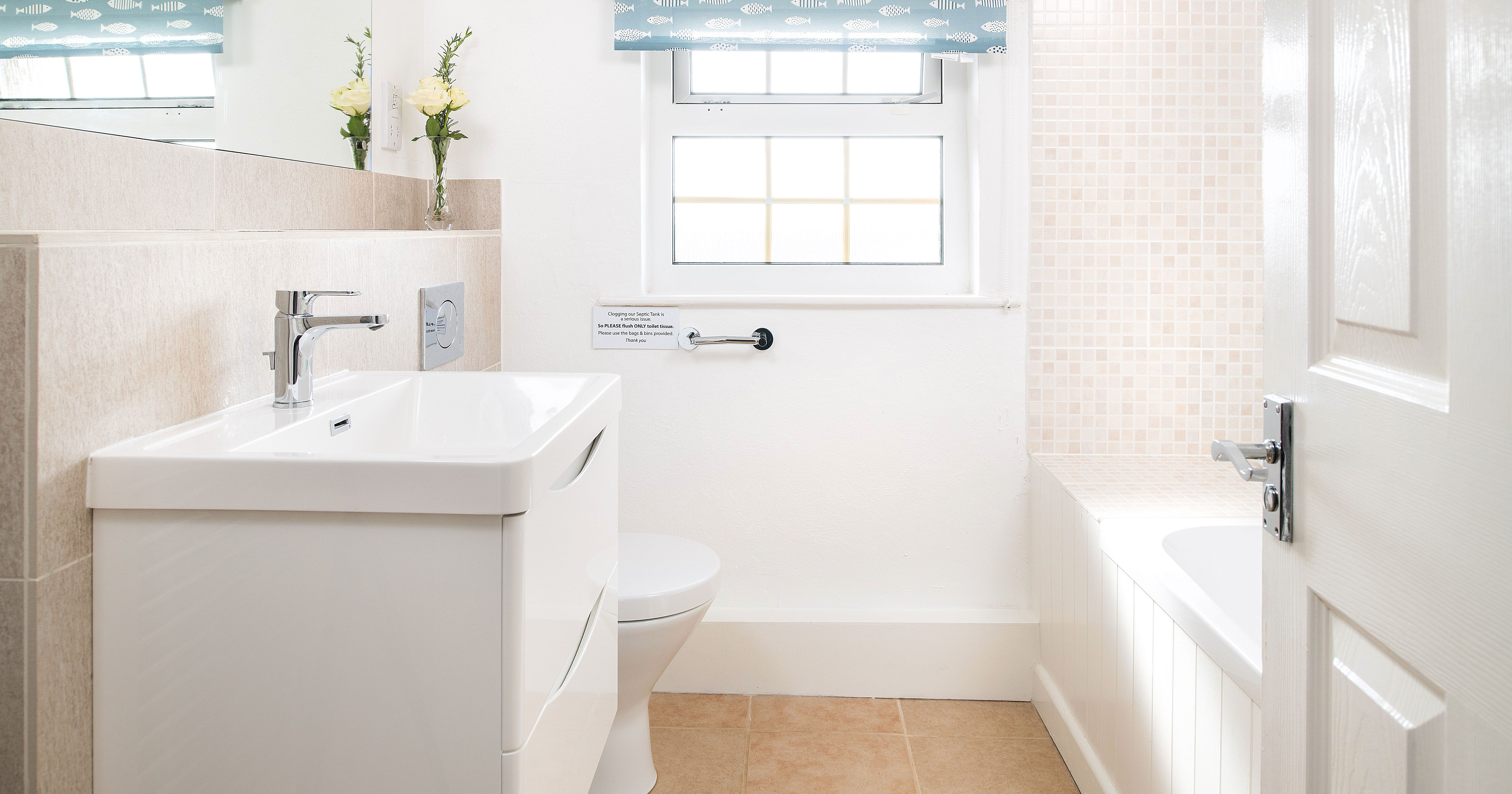 Bright, modern bathroom with white sink, toilet, and bathtub, beige tiles, a window with a blue patterned blind, and a vase with yellow flowers on the sink counter.