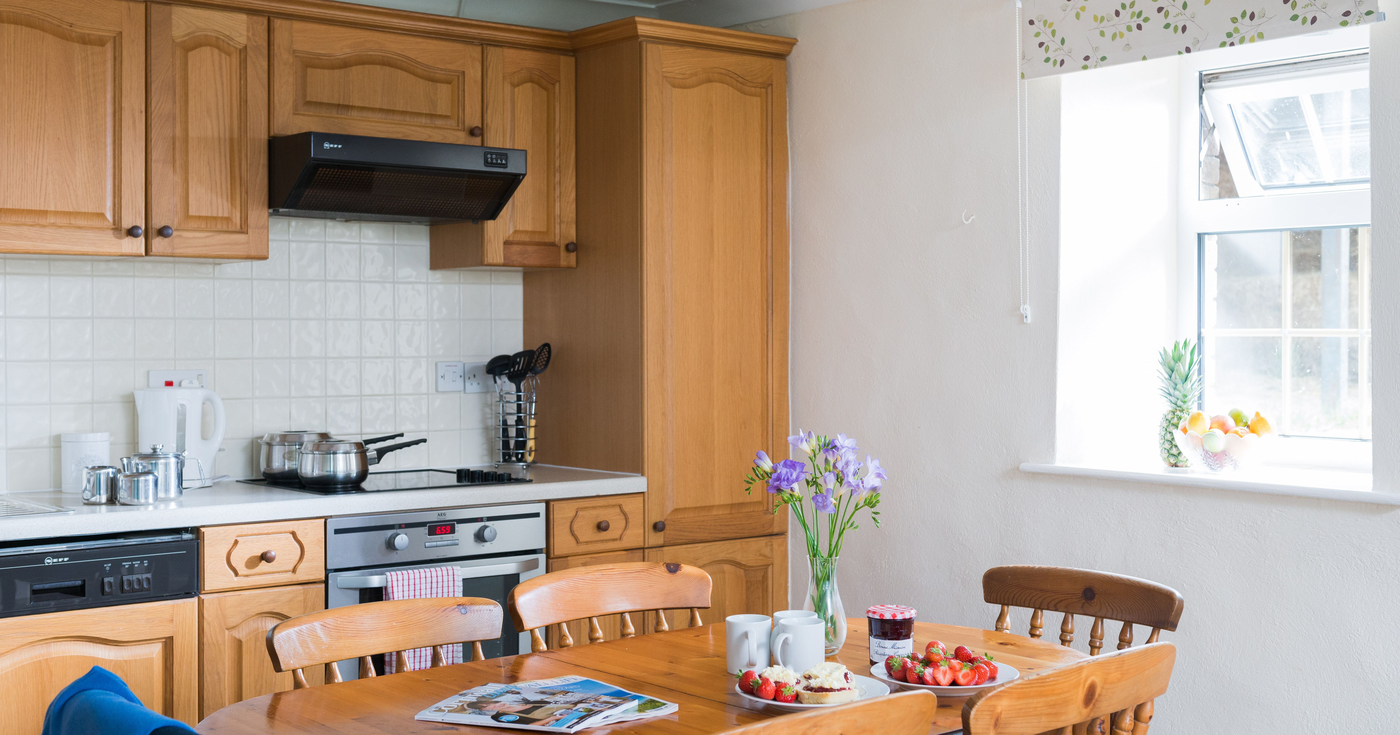 Cozy kitchen with wooden cabinets, a table set with strawberries, scones, jam, flowers, and mugs. Sunlit window with fruit bowl and pineapple on the sill.