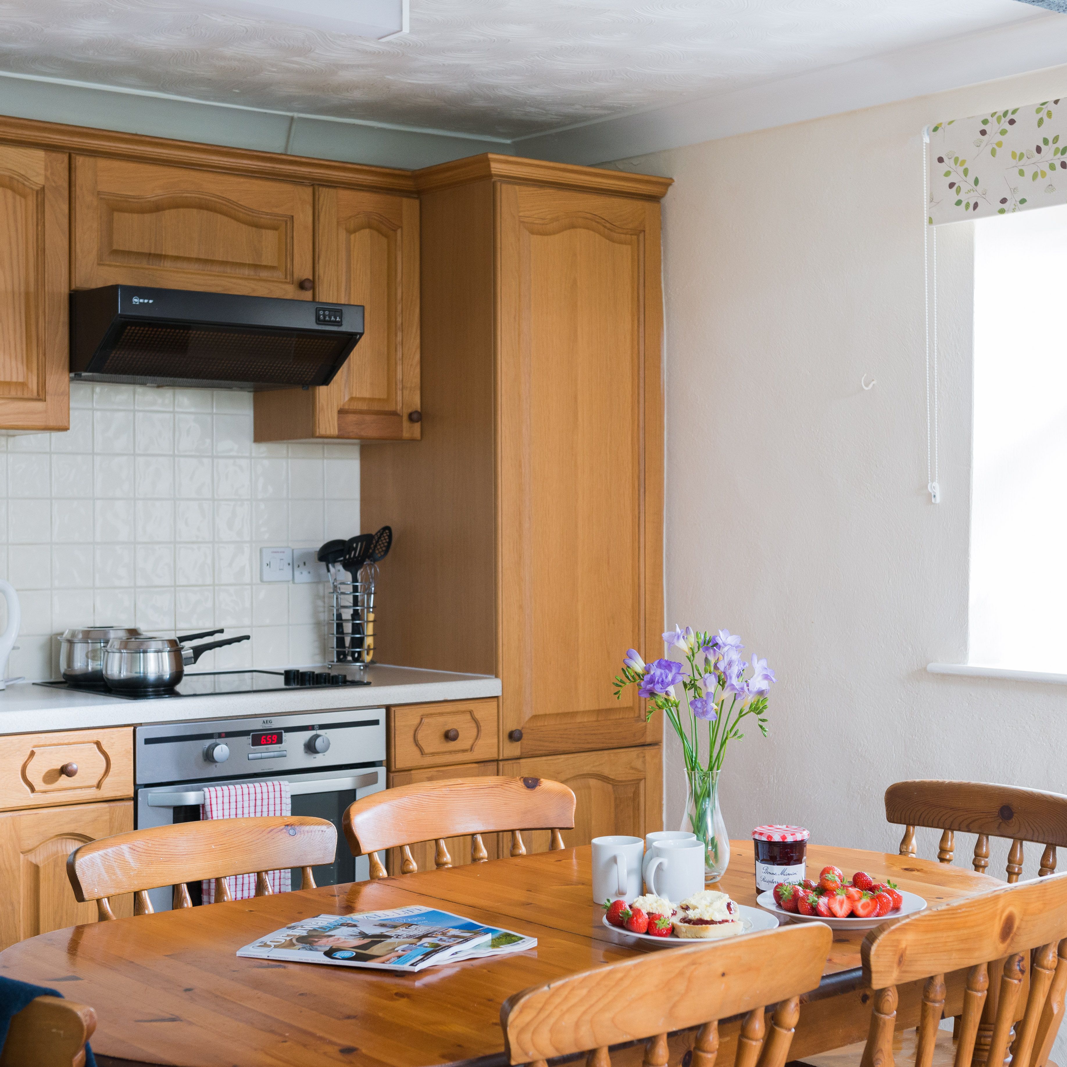 Cozy kitchen with wooden cabinets, a table set with strawberries, scones, jam, flowers, and mugs. Sunlit window with fruit bowl and pineapple on the sill.