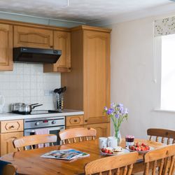 Cozy kitchen with wooden cabinets, a table set with strawberries, scones, jam, flowers, and mugs. Sunlit window with fruit bowl and pineapple on the sill.