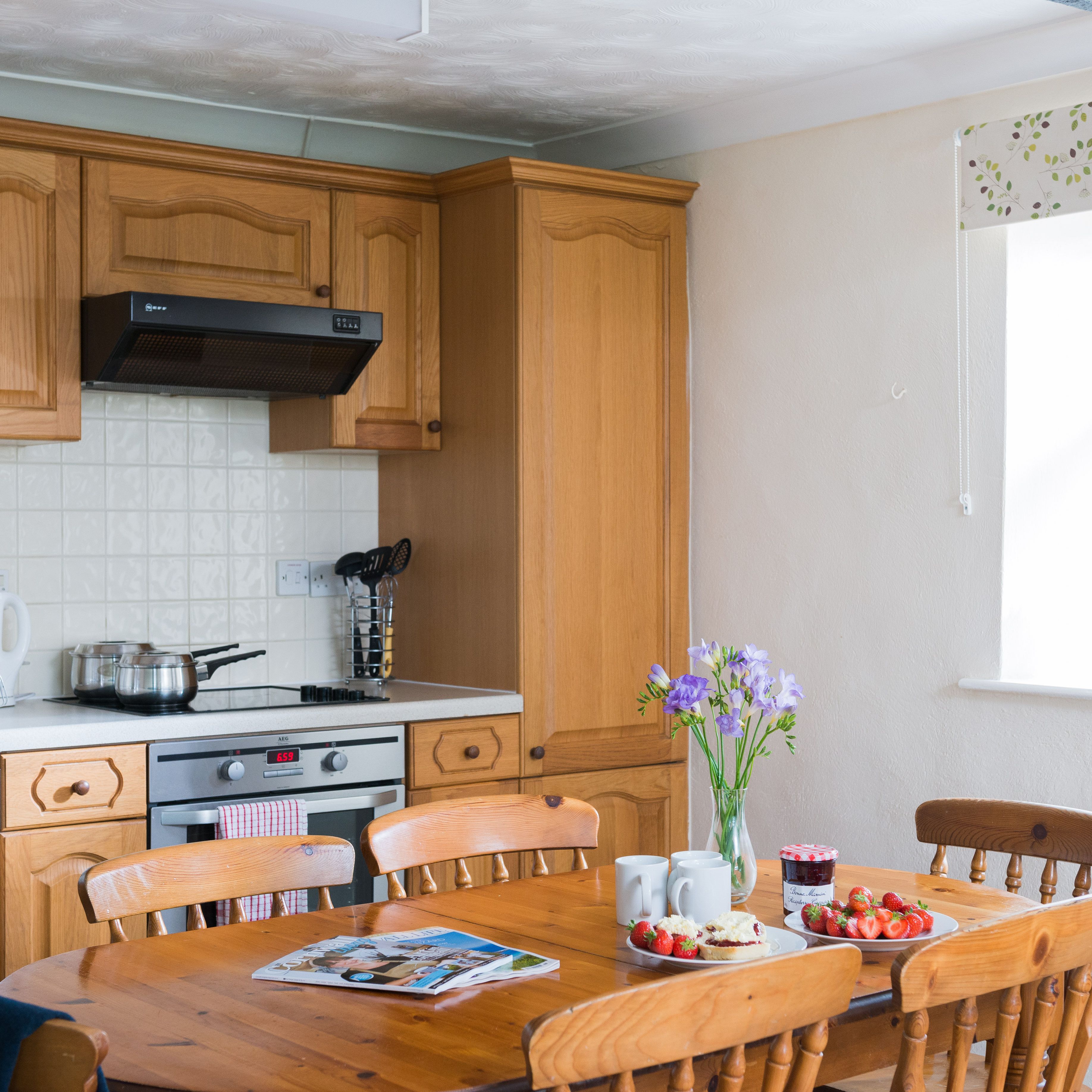Cozy kitchen with wooden cabinets, a table set with strawberries, scones, jam, flowers, and mugs. Sunlit window with fruit bowl and pineapple on the sill.